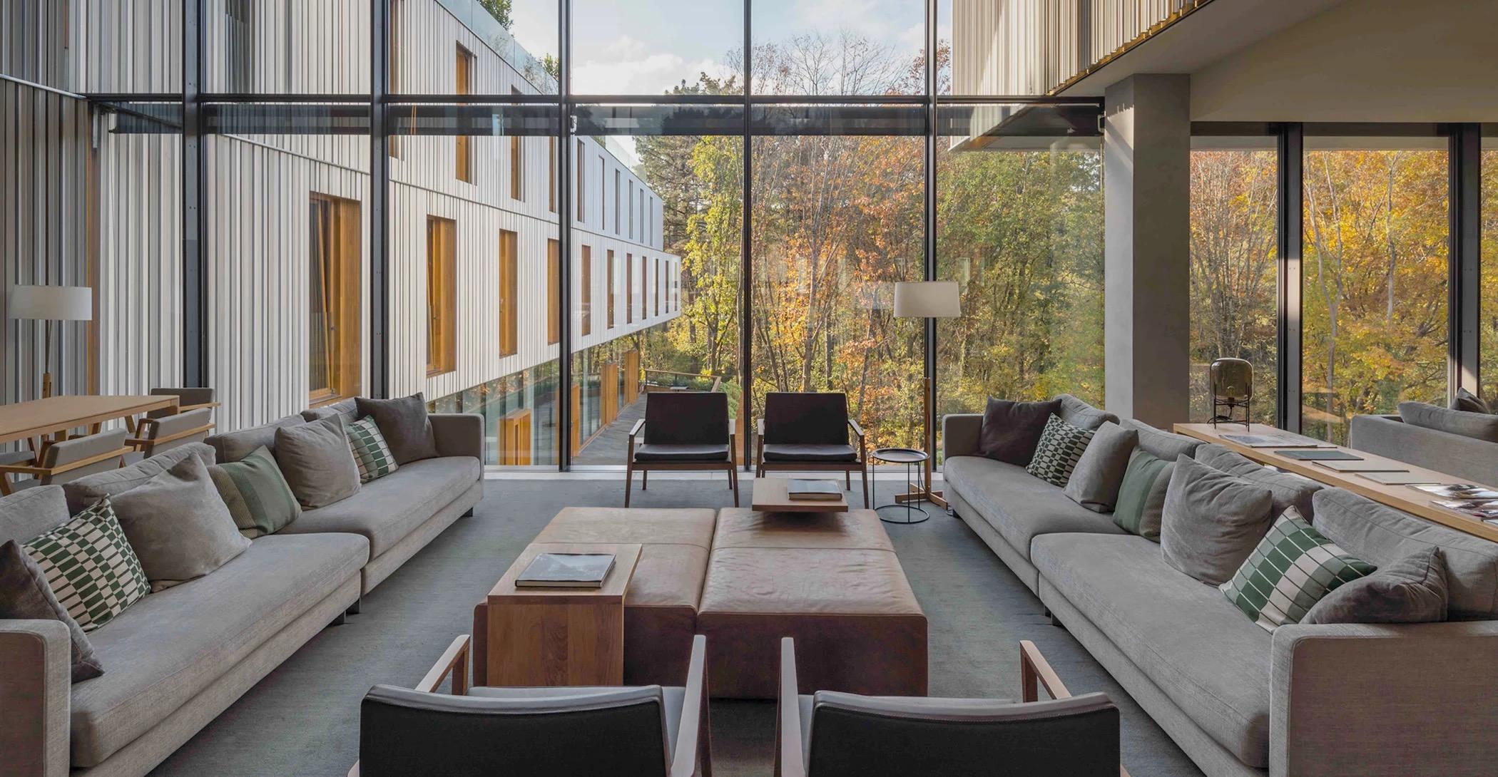 A double-height hotel lounge with two grey sofas facing each other, floor-to-ceiling windows looking onto the building's timber-clad facade and autumn trees.