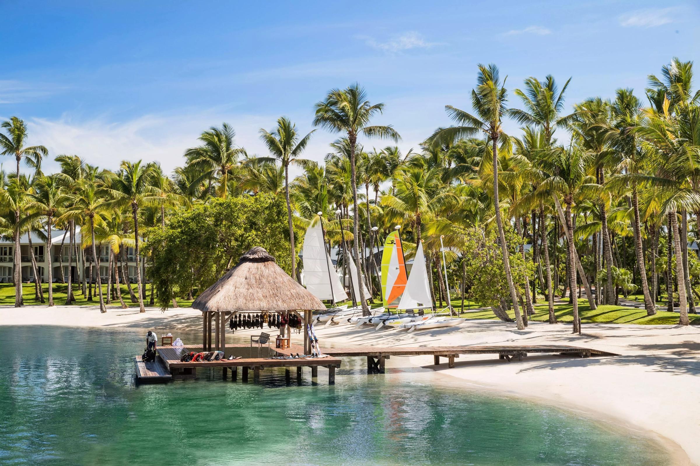 A thatched hut on a wooden jetty holds sailing gear, with small catamarans pulled up on the sand and palm trees and a building behind.