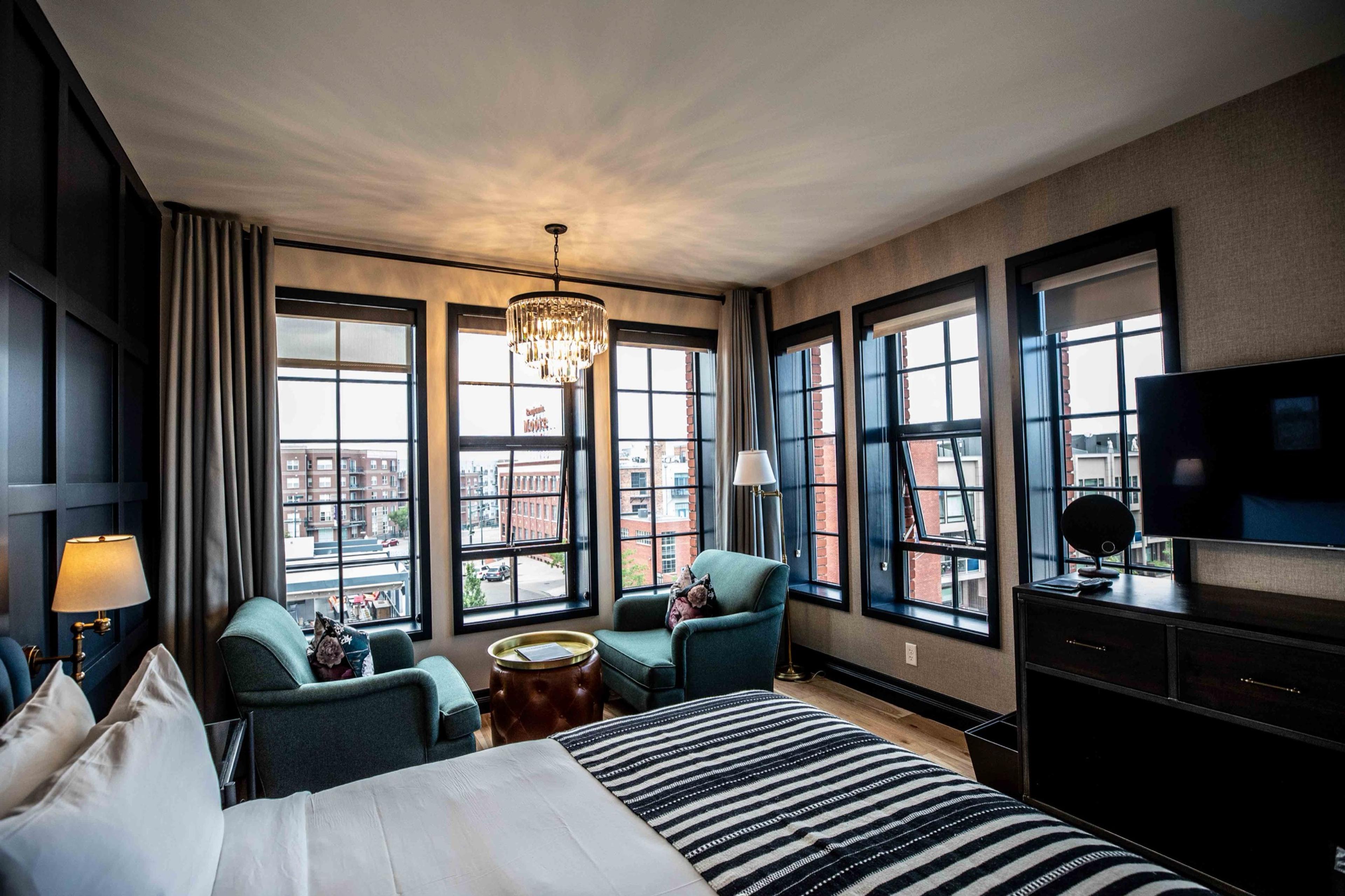 A corner room with two teal armchairs by a wall of windows overlooking the city, a chandelier, and a television on a dark dresser.