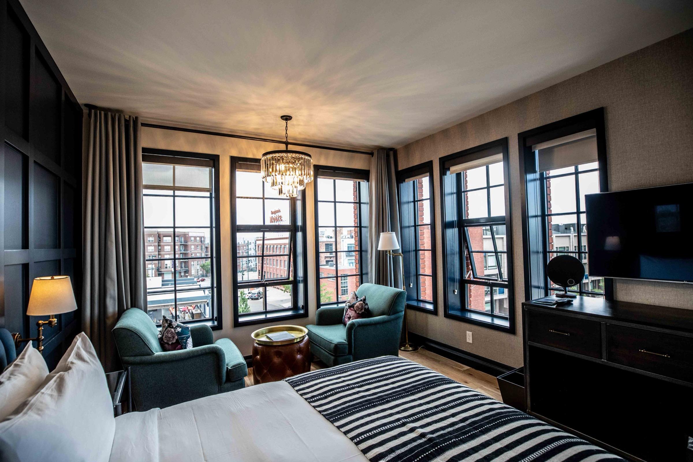 A corner room with two teal armchairs by a wall of windows overlooking the city, a chandelier, and a television on a dark dresser.