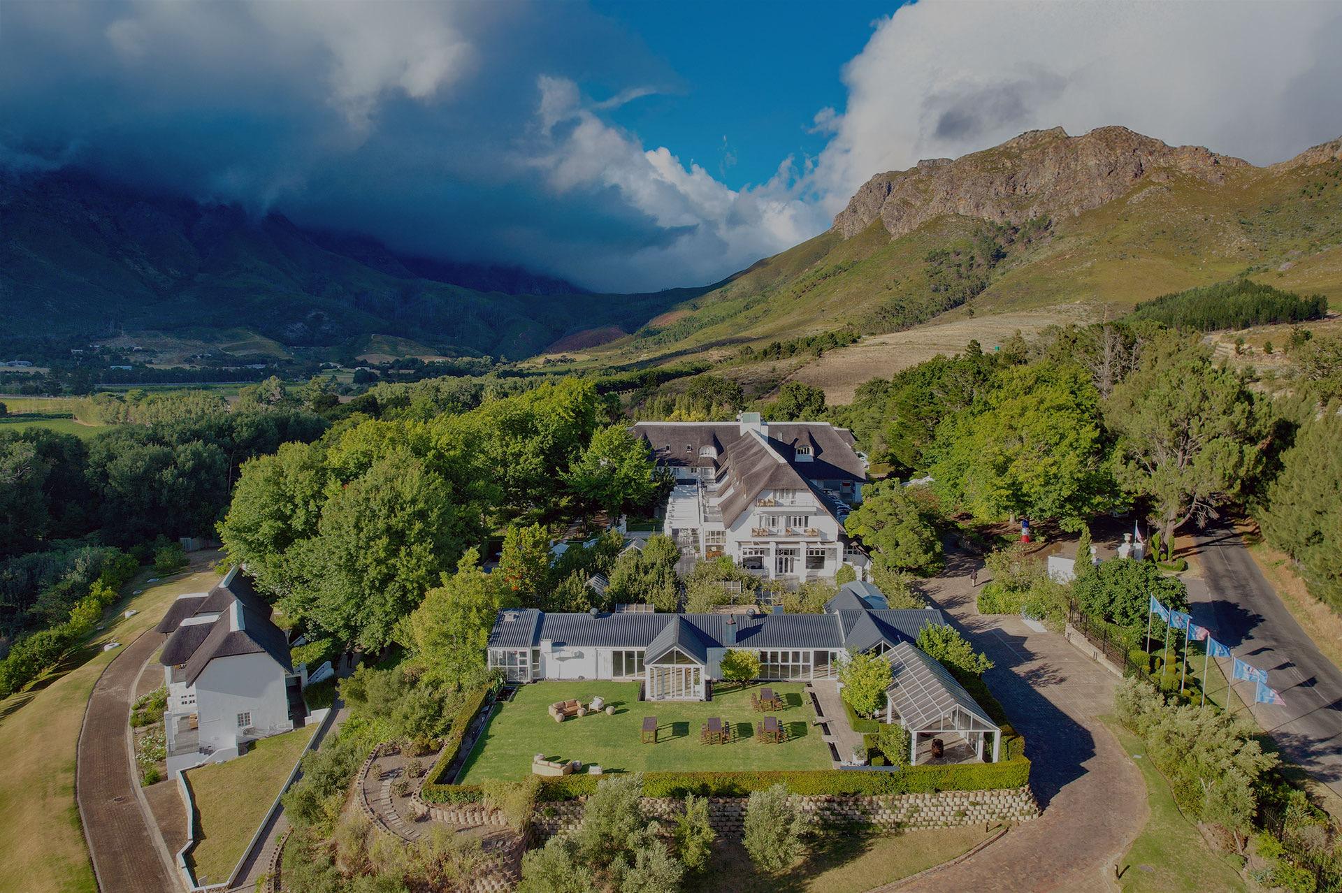 An aerial view of a white hotel with a thatched roof, a lawn with chairs and a glass-roofed pavilion, set among trees with mountains behind.
