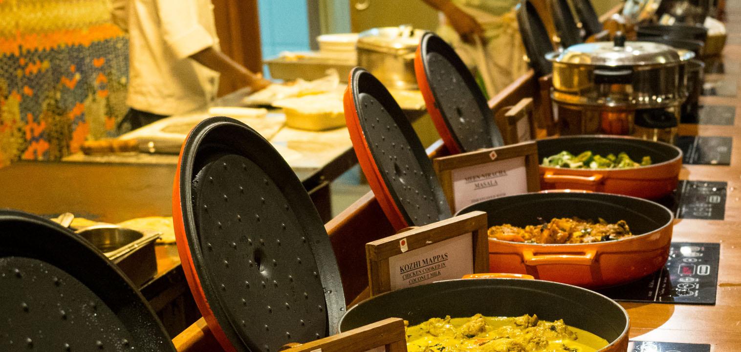 A row of open buffet dishes holding curries and vegetables, each labelled on a small card, with blurred figures behind.