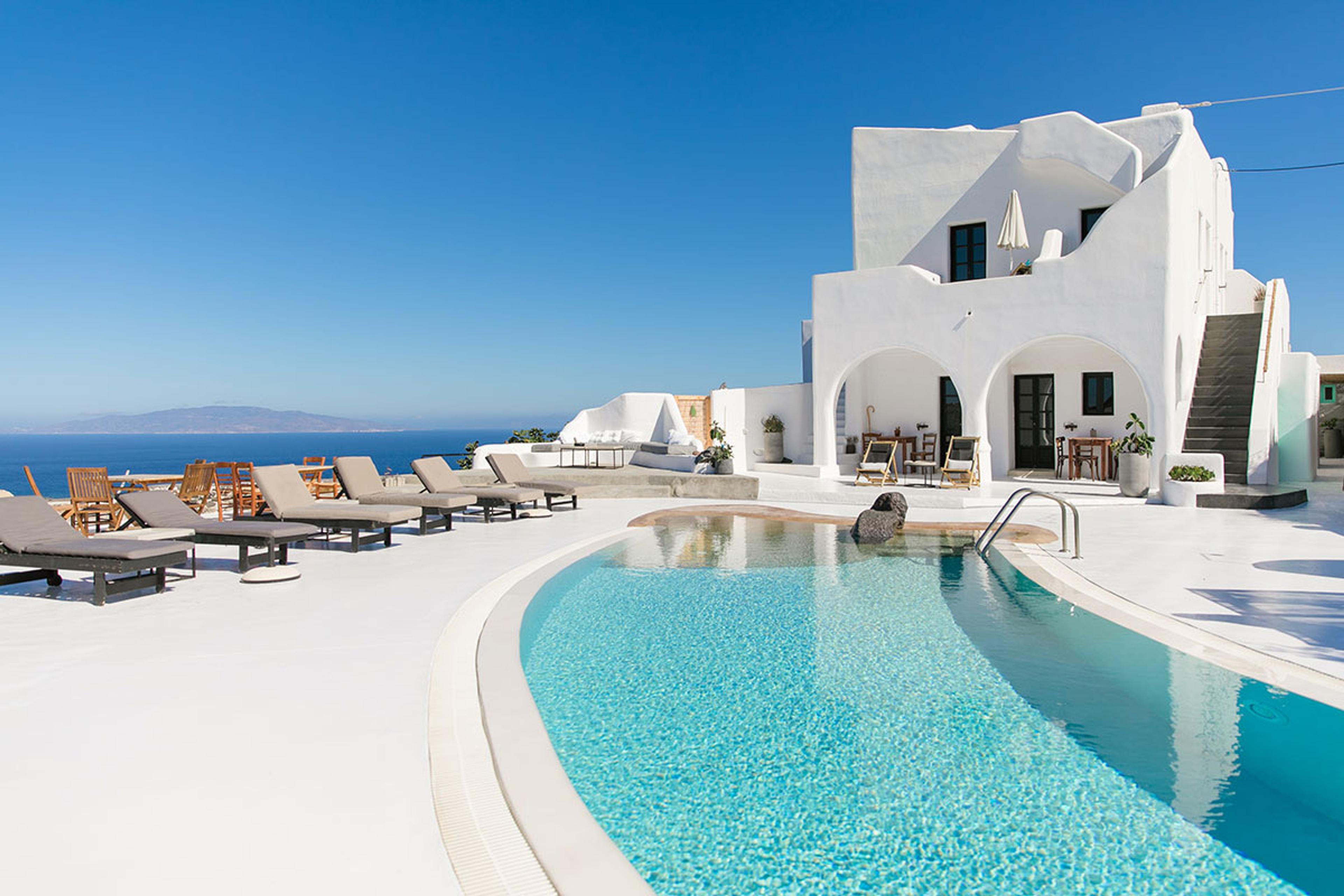 A white terrace beside a turquoise pool, with pale loungers and parasols on the left, a two-storey white building with arched doorways behind, and the sea and a distant island under a clear blue sky.