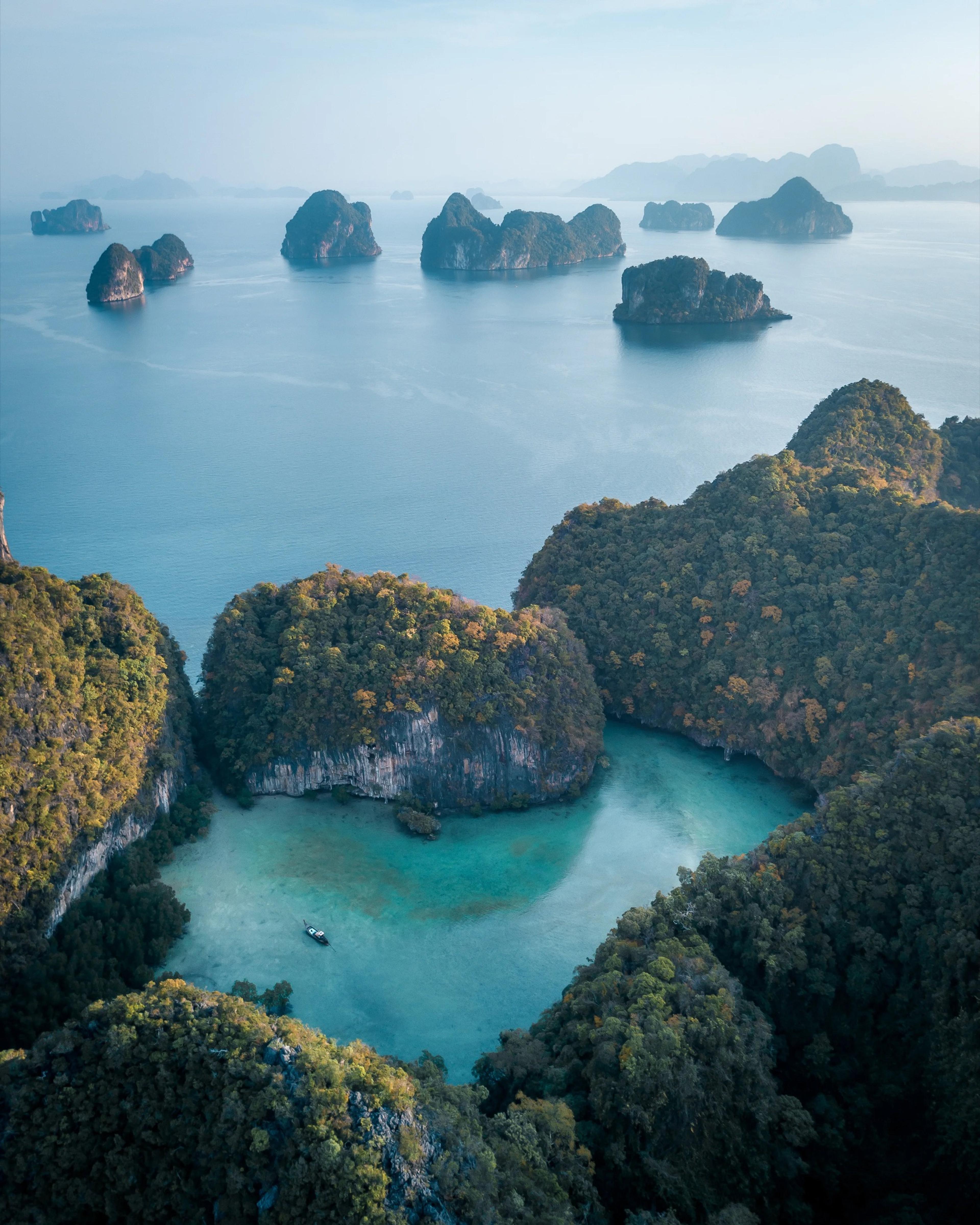 An aerial view of forested limestone islands rising from a clear turquoise bay, with more islets scattered across the water towards a hazy horizon.