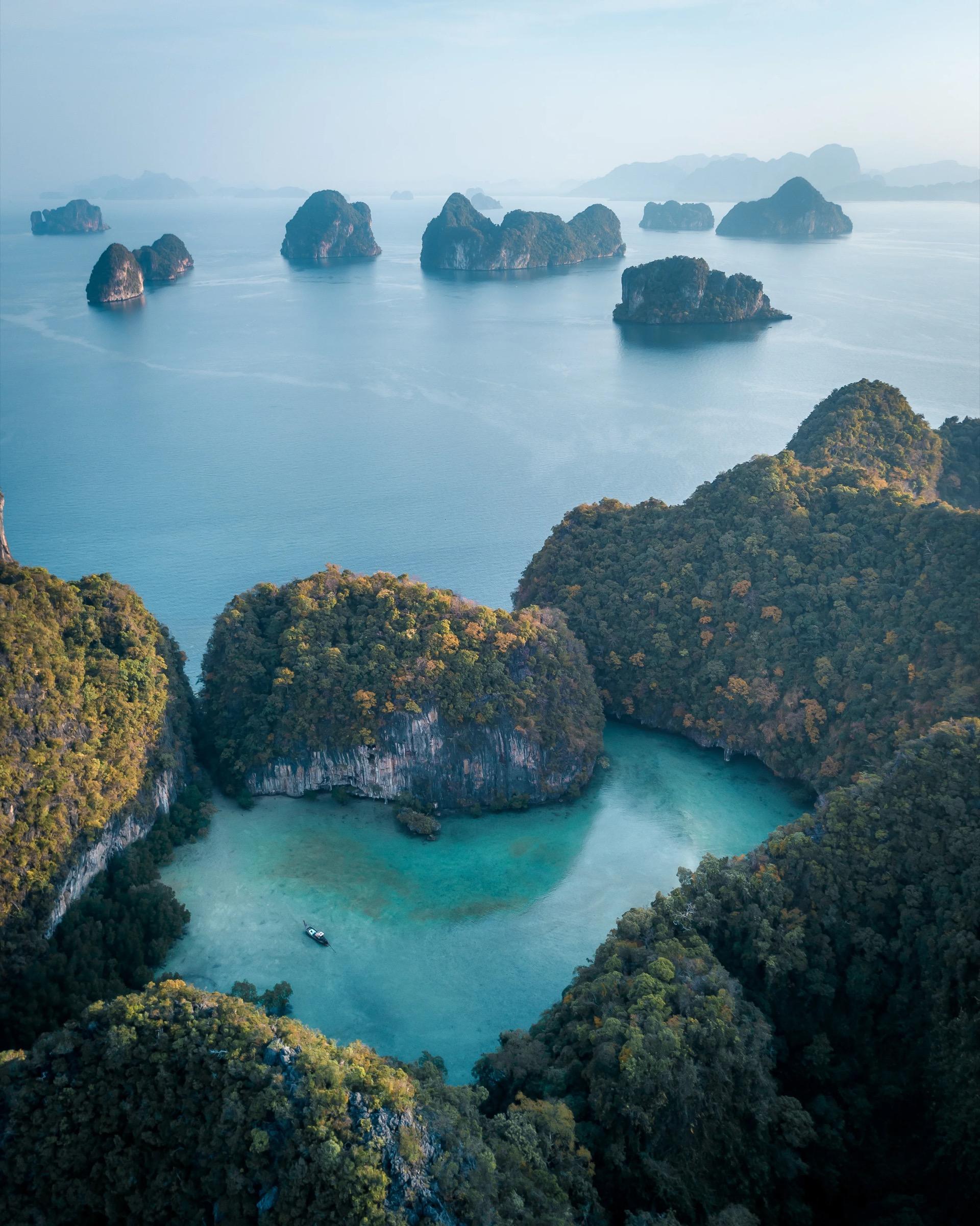 An aerial view of forested limestone islands rising from a clear turquoise bay, with more islets scattered across the water towards a hazy horizon.