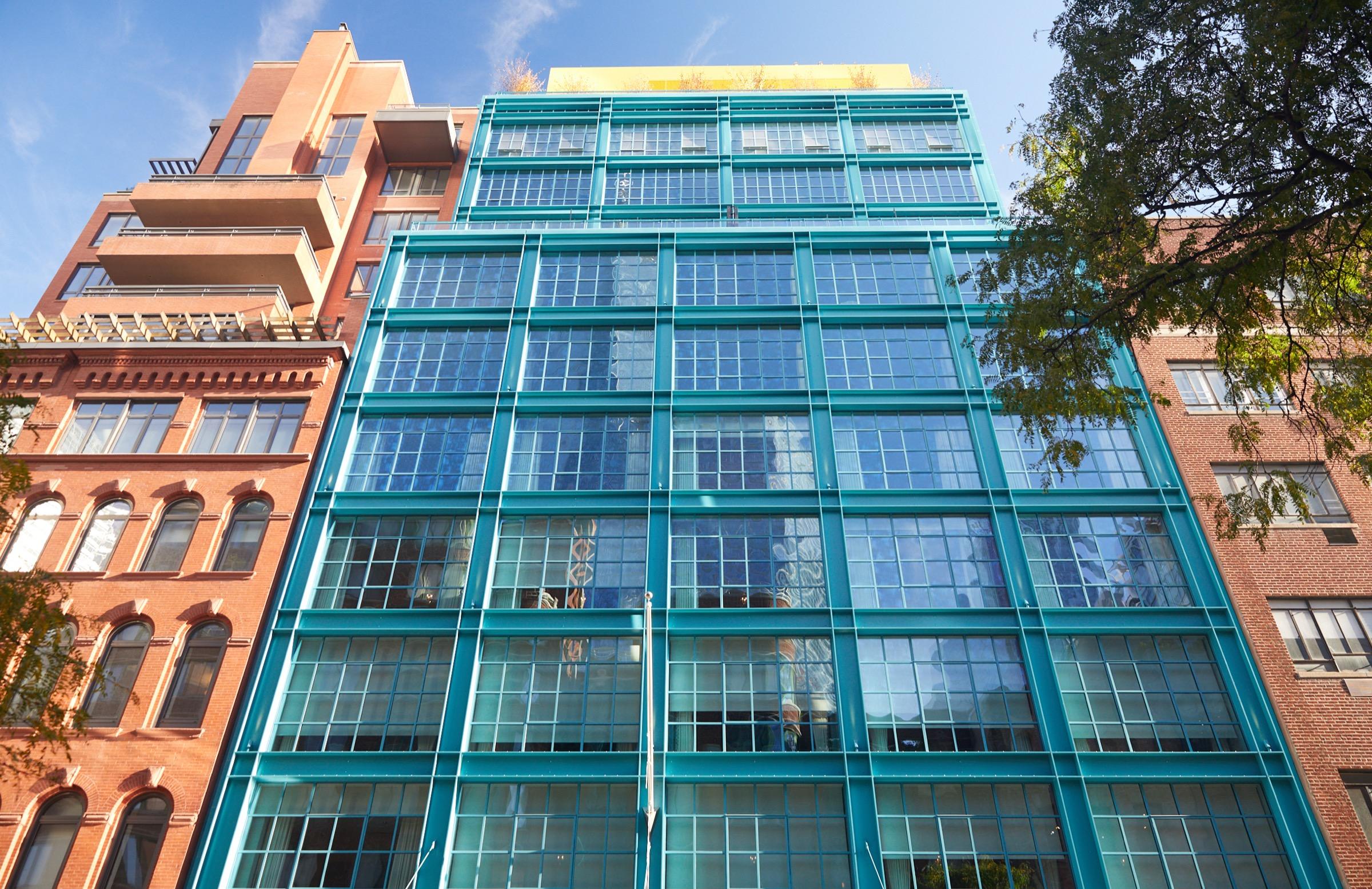A teal glass and steel building rising above its red-brick neighbours, photographed from street level looking up against a blue sky.