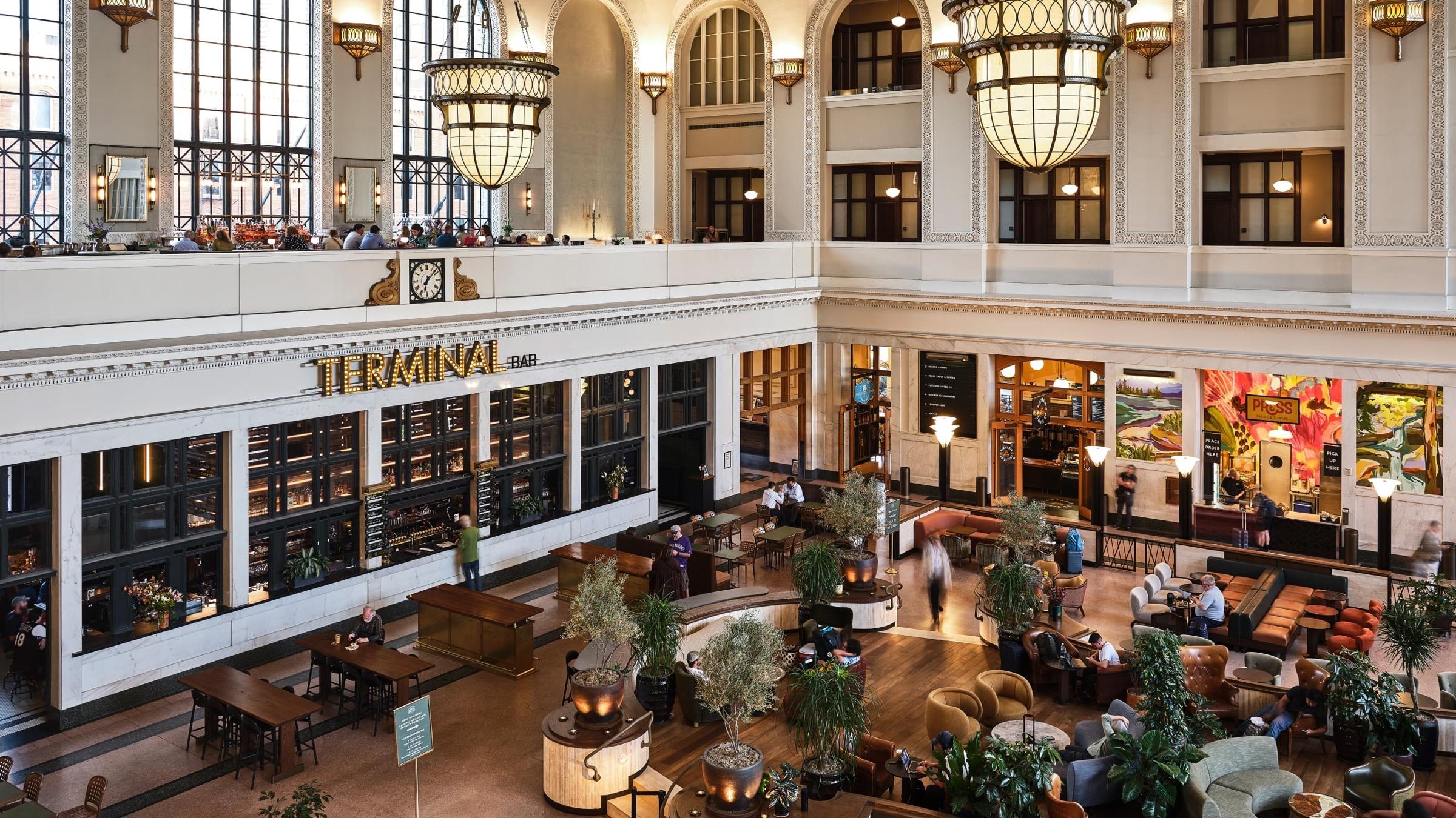 A tall arched hall seen from an upper level, with a chandelier, a bar labelled Terminal Bar, a balcony with people along its rail, and seating on the floor below.