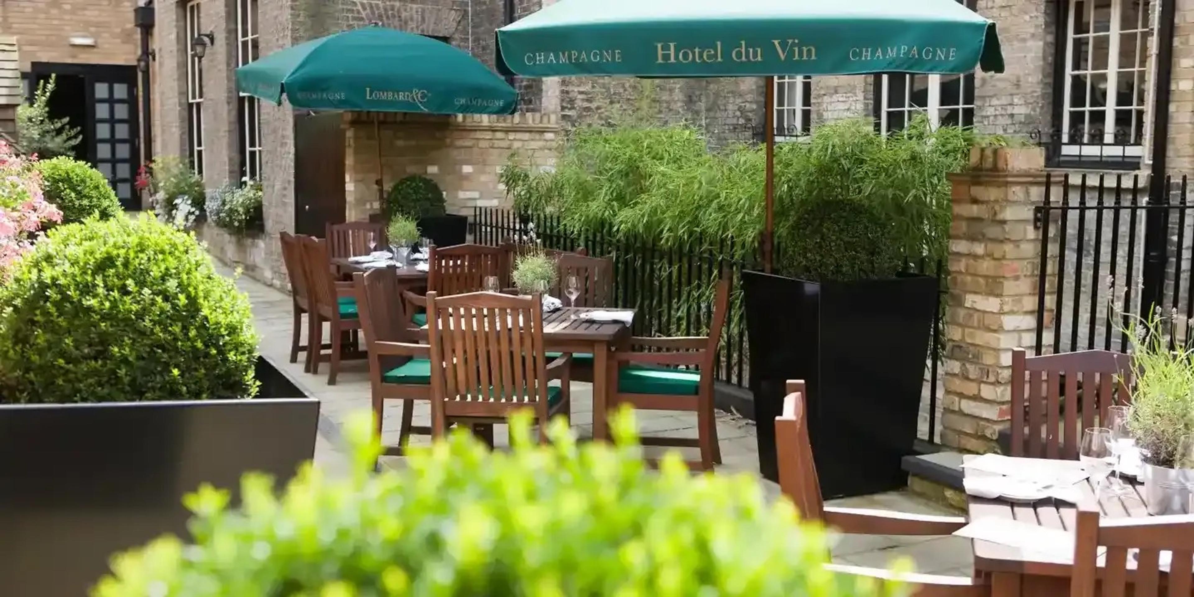A paved courtyard patio beside old brick buildings, with green parasols, wooden chairs with green seat pads at tables, and clipped box hedges in black planters.