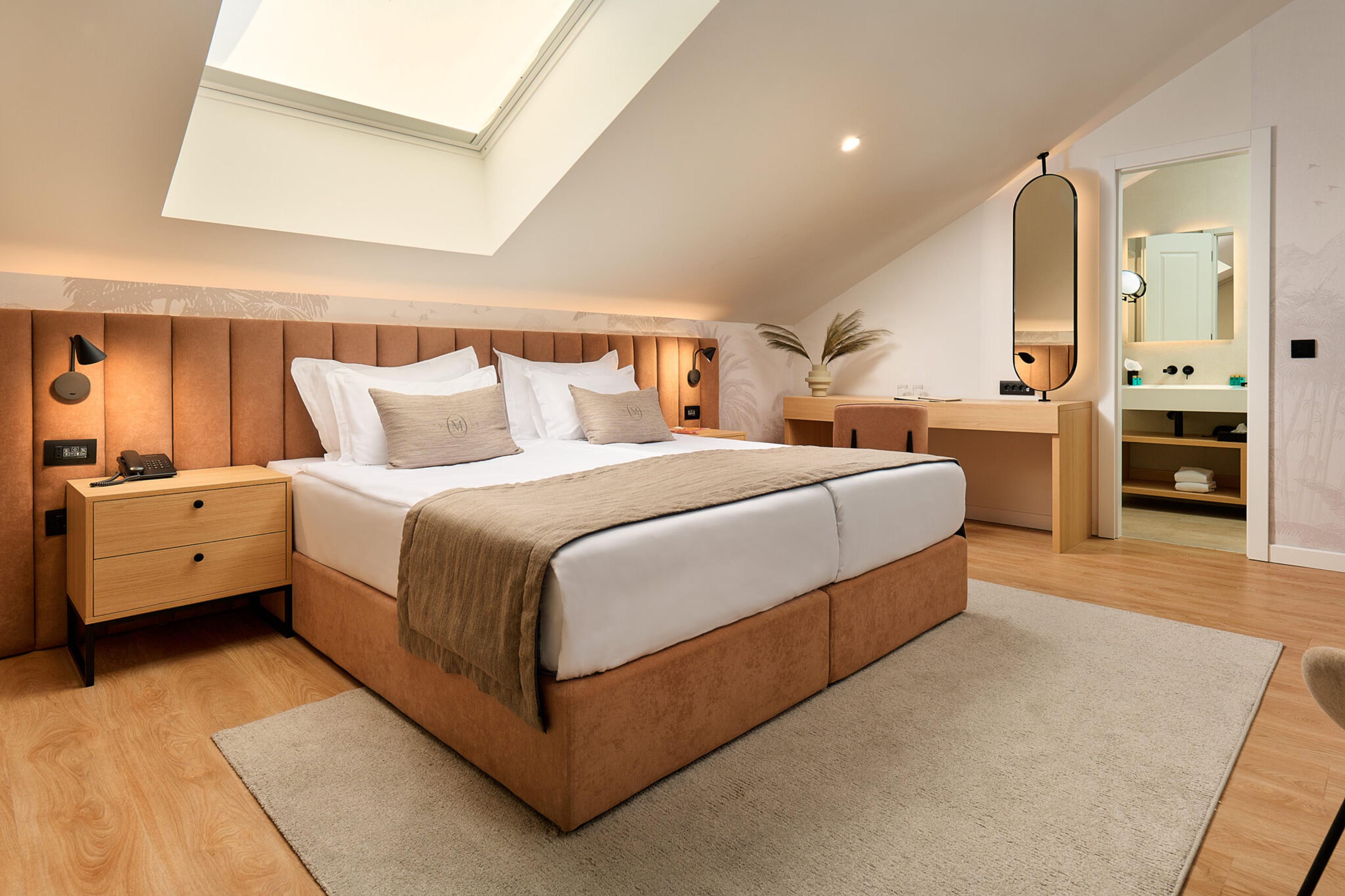 A bedroom under a sloped skylight, with a tan upholstered bed, a wooden desk and an open door to an ensuite bathroom.
