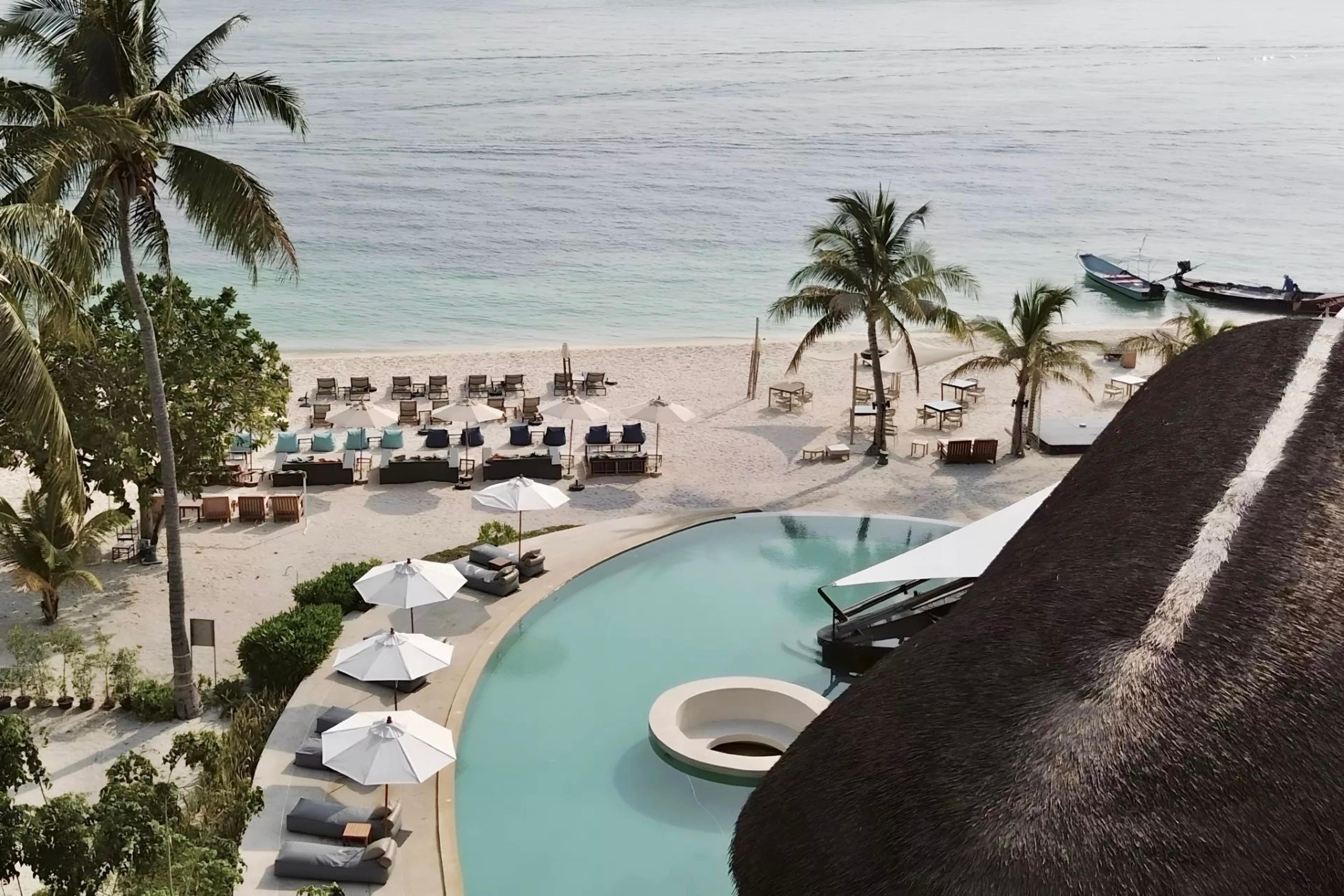 a view from a balcony over an infinity pool to a white sand beach with sunbeds, palm trees, and boats moored offshore