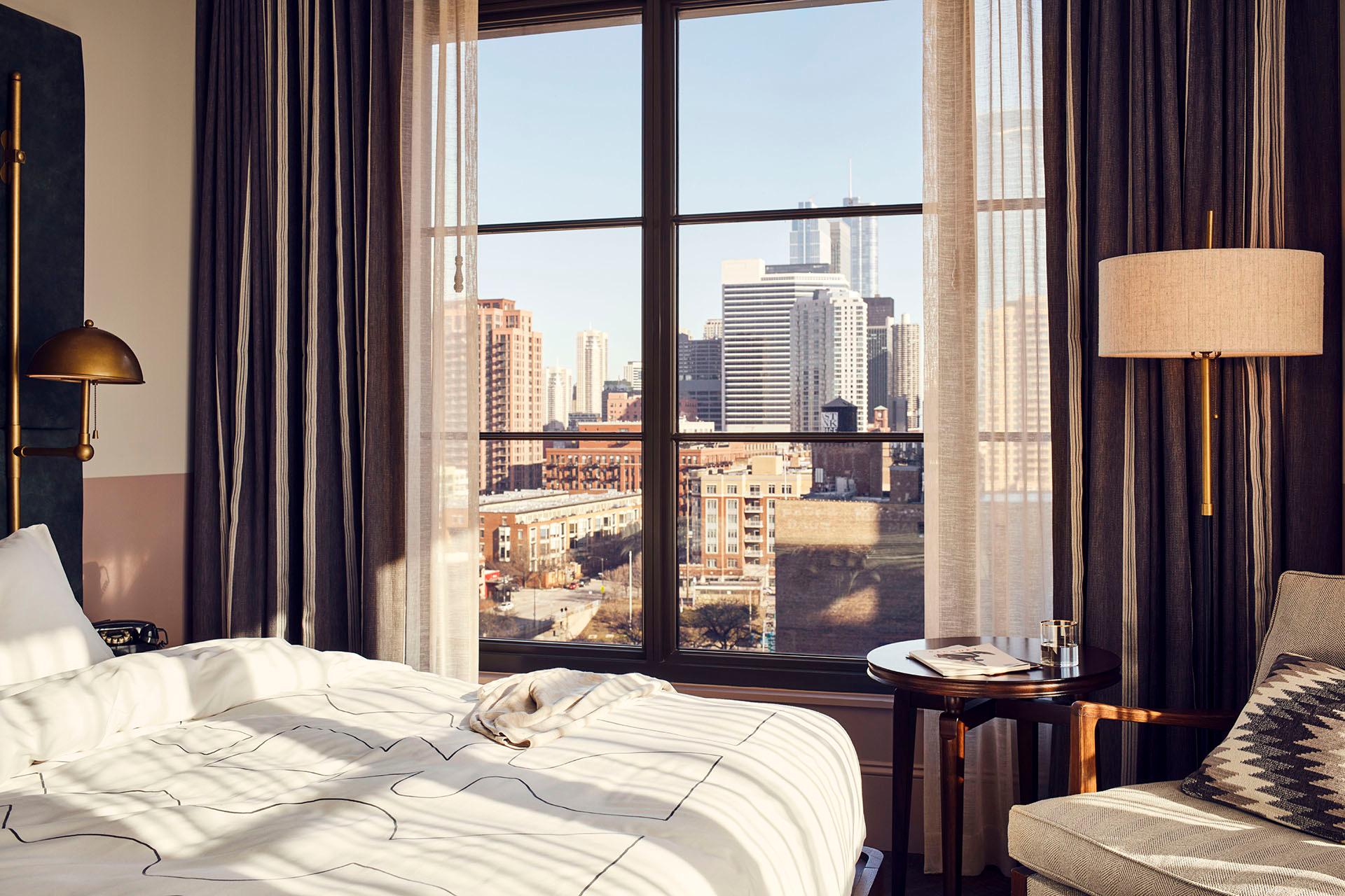 A hotel bed with a grey tufted headboard in front of a large window showing the Chicago skyline in daylight, with a round side table and armchair alongside.