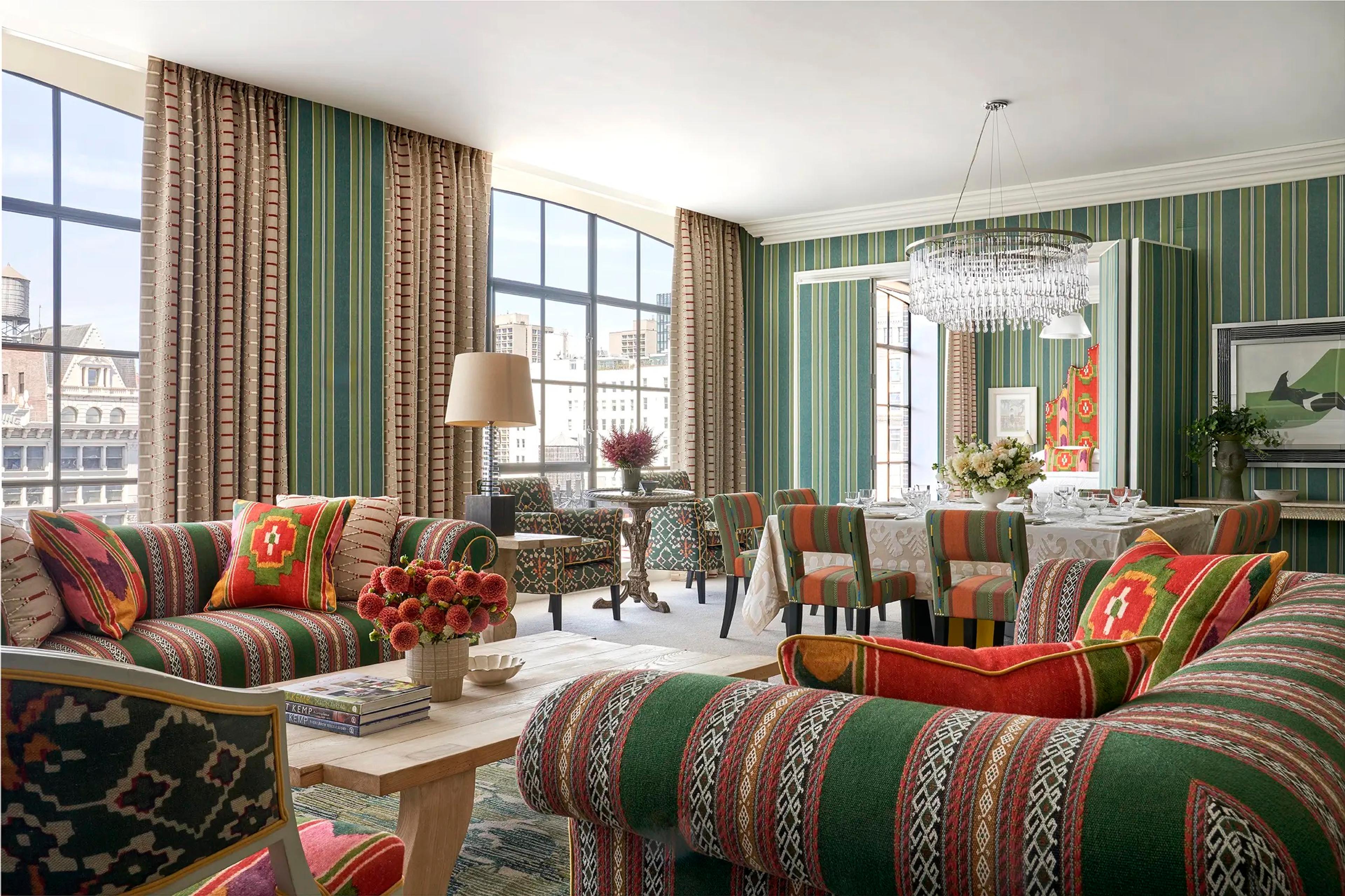 A suite sitting and dining room with green striped wallpaper, striped sofas, a dining table set for a meal, and city rooftops with a water tower visible through tall windows.