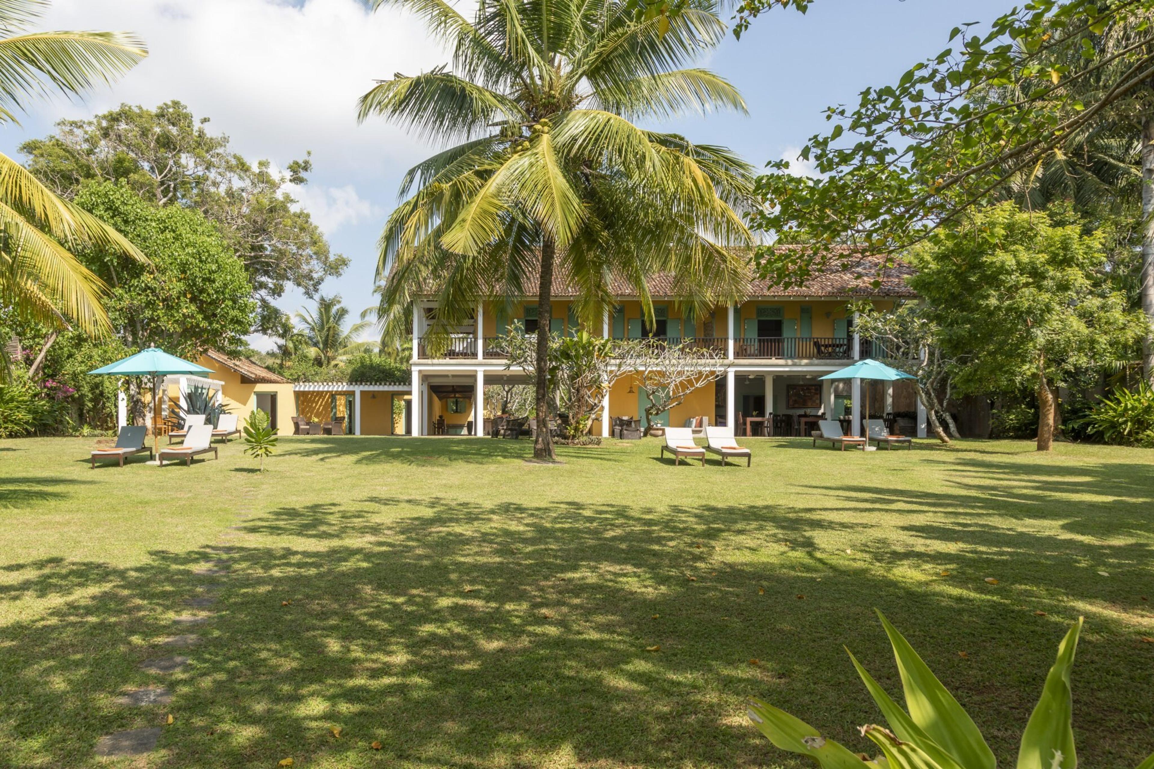A two storey yellow house with wraparound verandas and balconies stands behind a wide lawn with sun loungers, blue umbrellas and a large palm tree, under a partly cloudy sky.