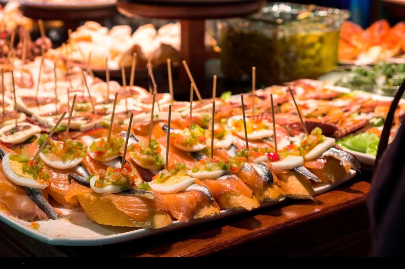 A platter of open sandwiches topped with anchovies, salmon and peppers, each pinned with a cocktail stick, on a restaurant counter.