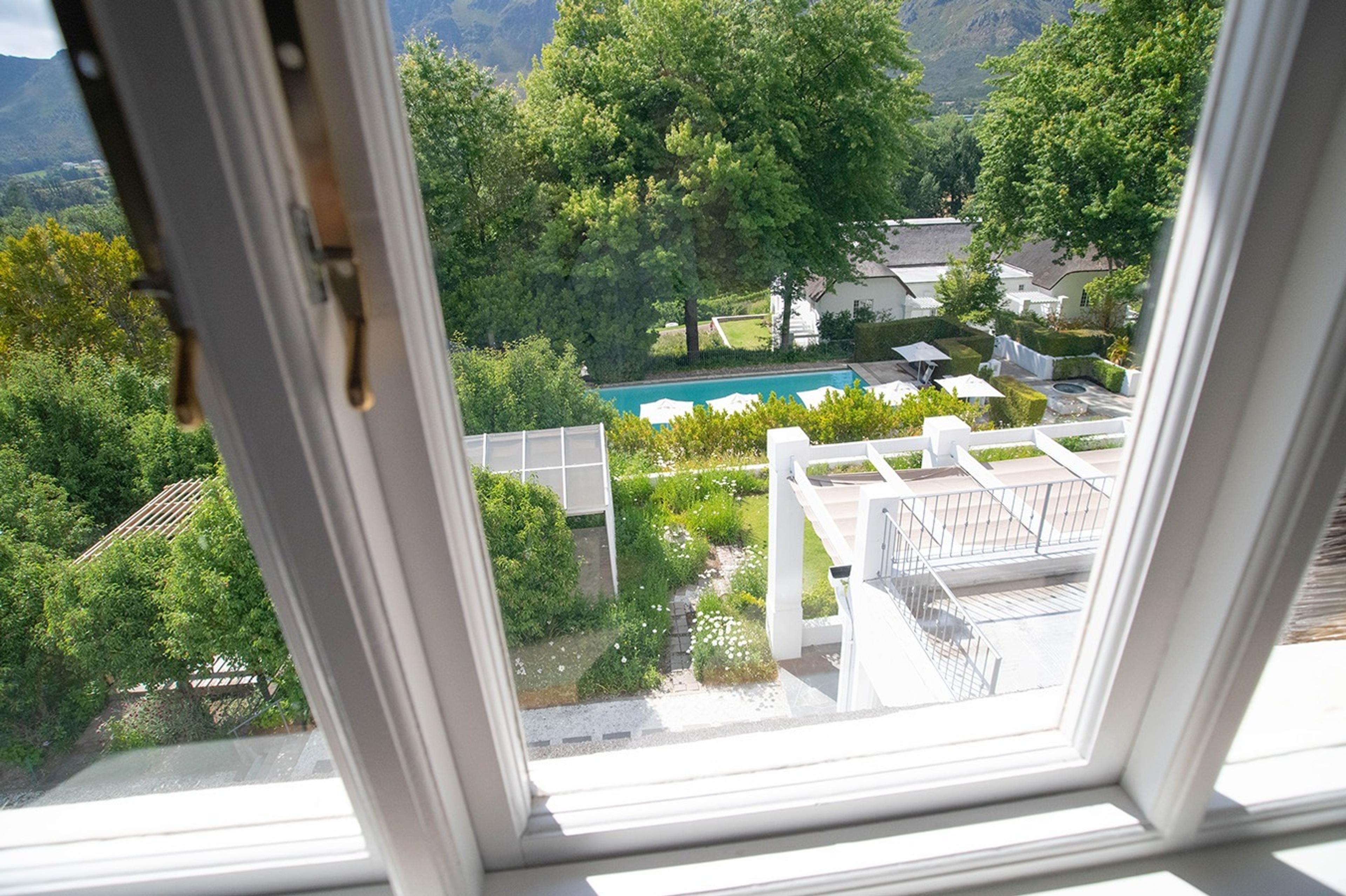 A pool and garden seen through a white window frame, with white parasols, a pergola and trees.