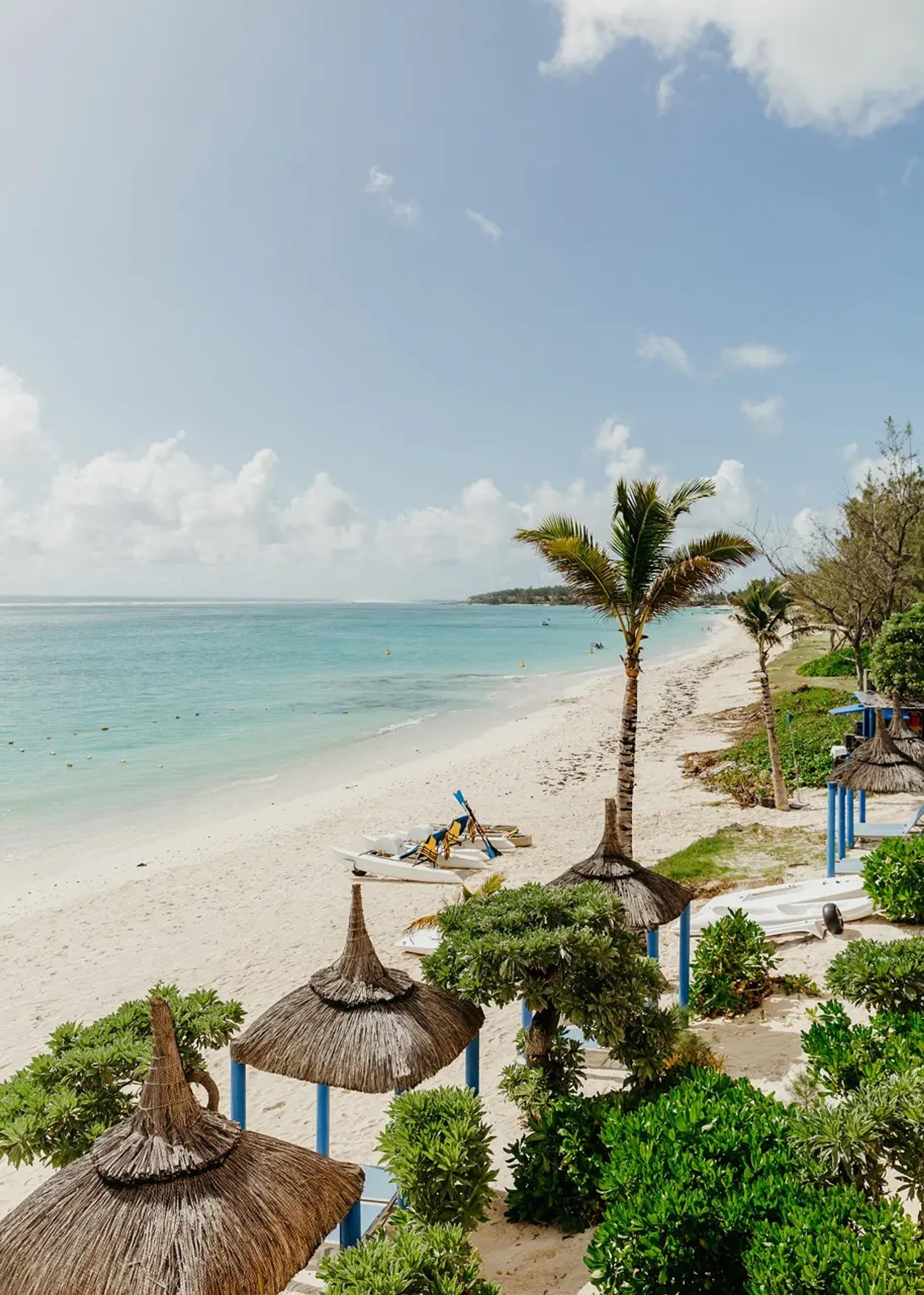 Thatched sunshades and a palm tree line a white sand beach beside a calm turquoise sea, with small boats pulled up on the sand.
