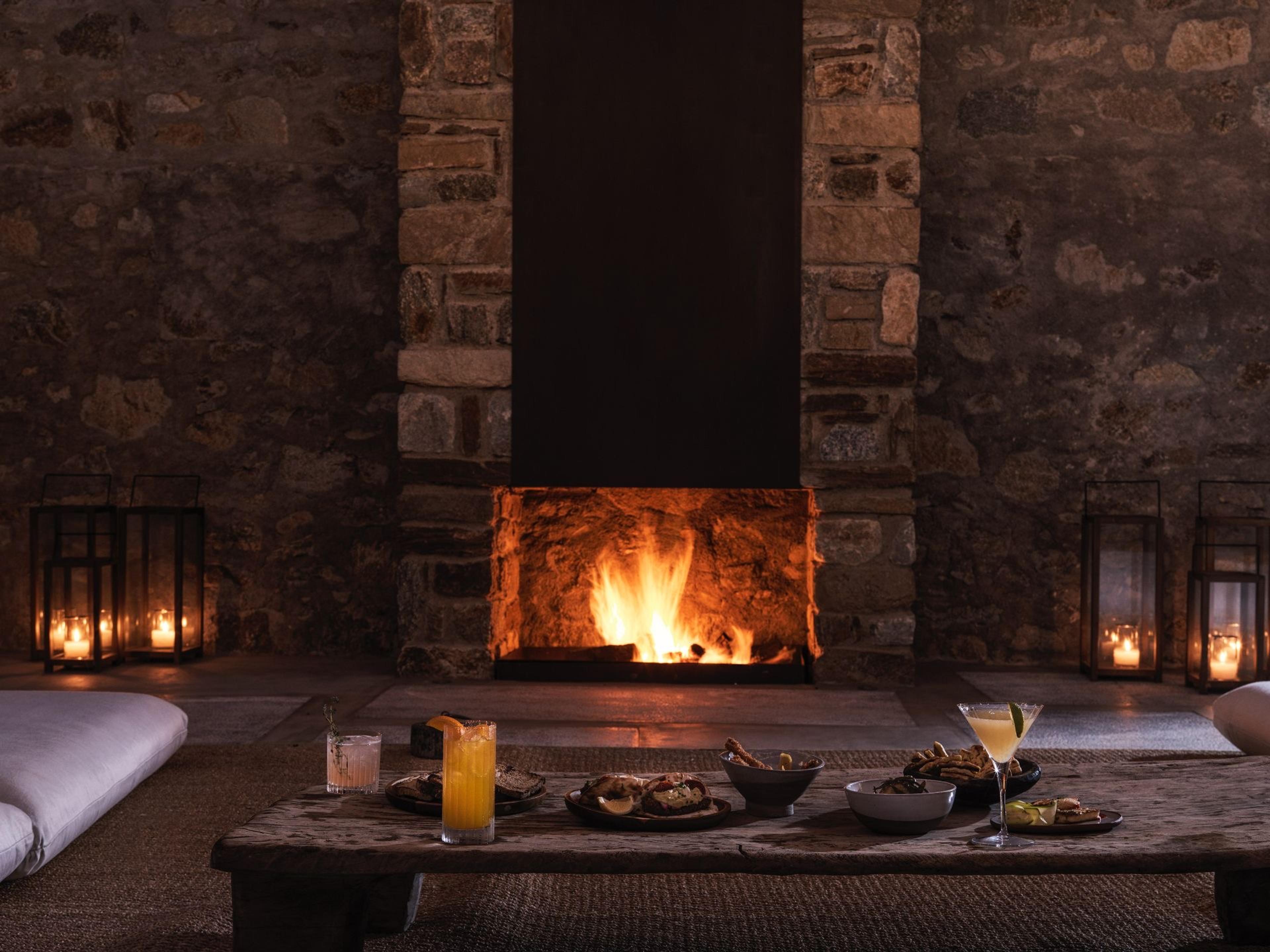 A low wooden table in front of a lit stone fireplace is set with cocktails, a glass of juice and small plates of sliders and dips, white cushions and black lanterns to either side.