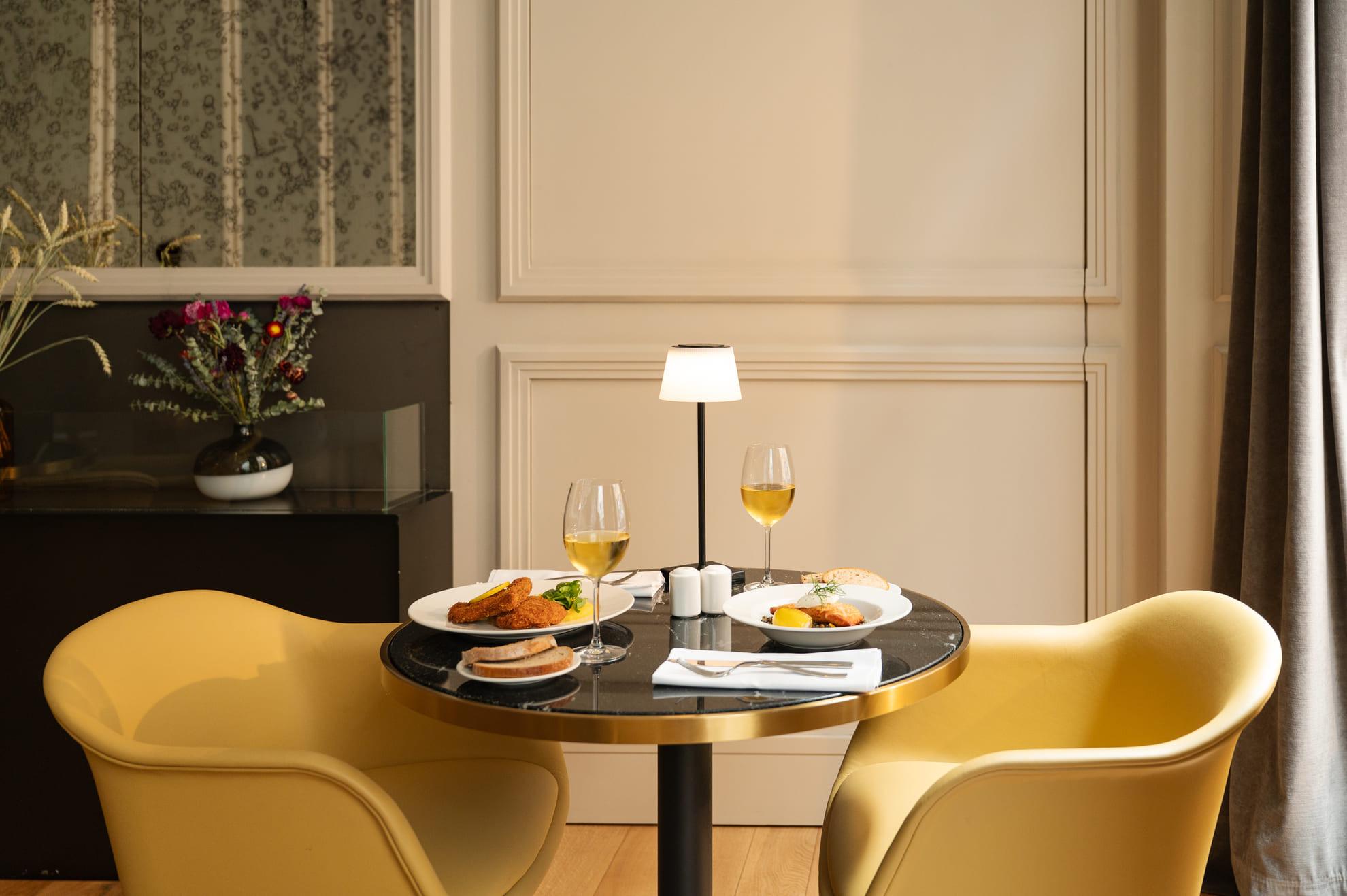 A round black-topped table with two plates of fried food and bread, and two glasses of white wine, between two yellow armchairs.