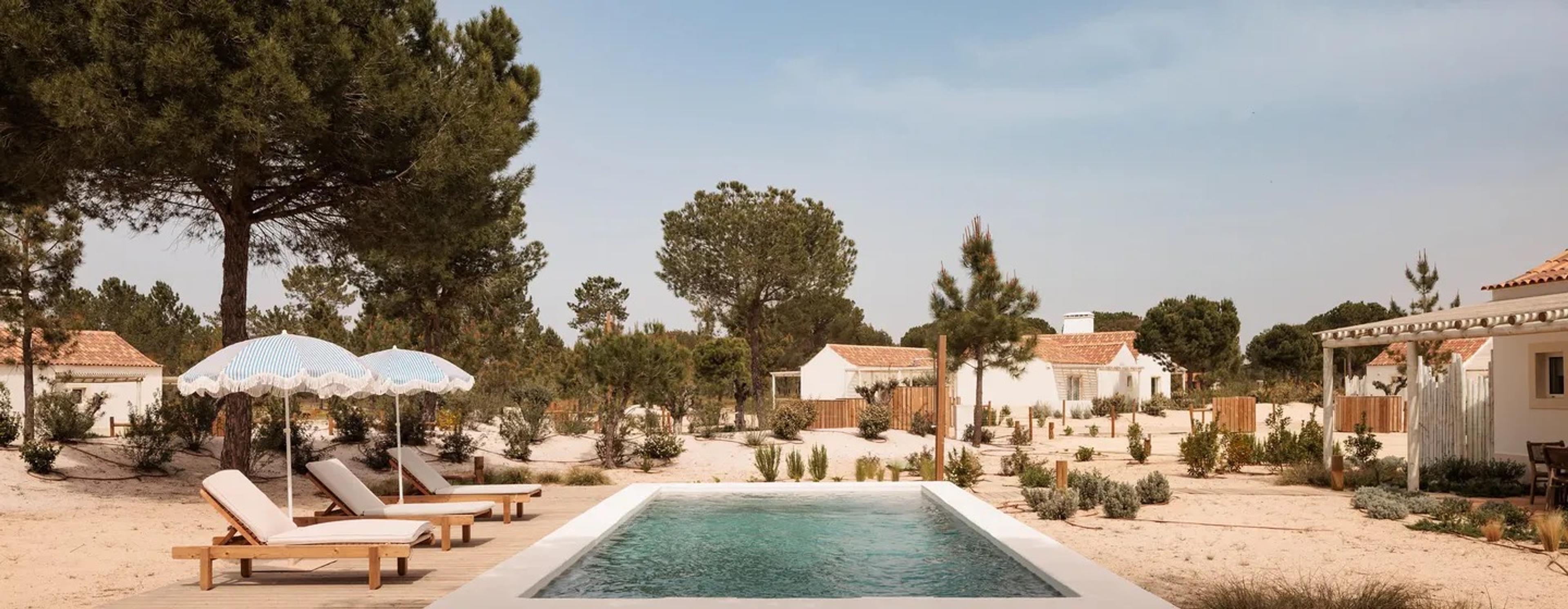 a swimming pool on a sandy terrace surrounded by white cottages with terracotta roofs and umbrella-shaded loungers, pine trees behind
