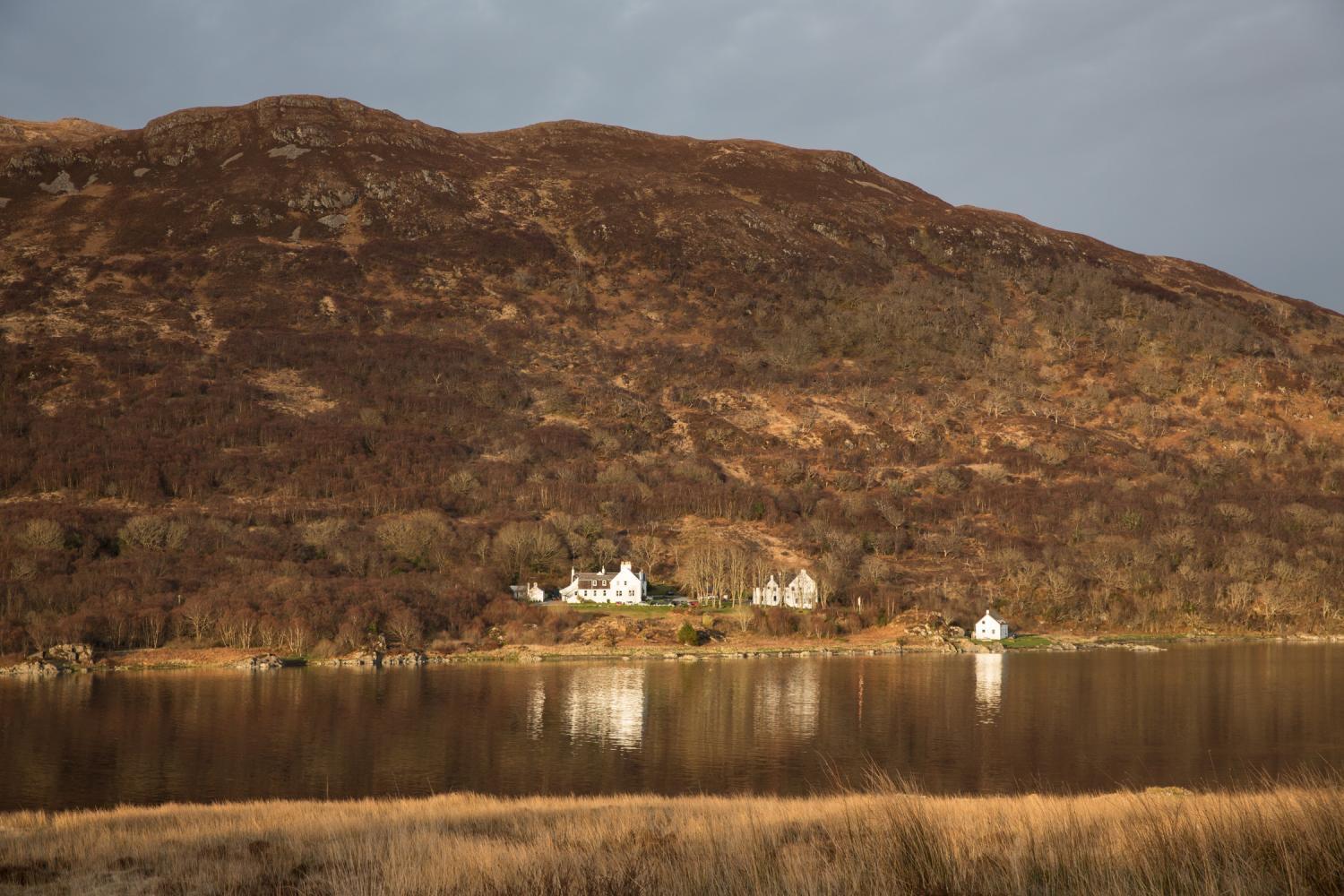 A white house and outbuildings on the far shore of a sea loch at low golden light, framed by a heather-covered hillside.