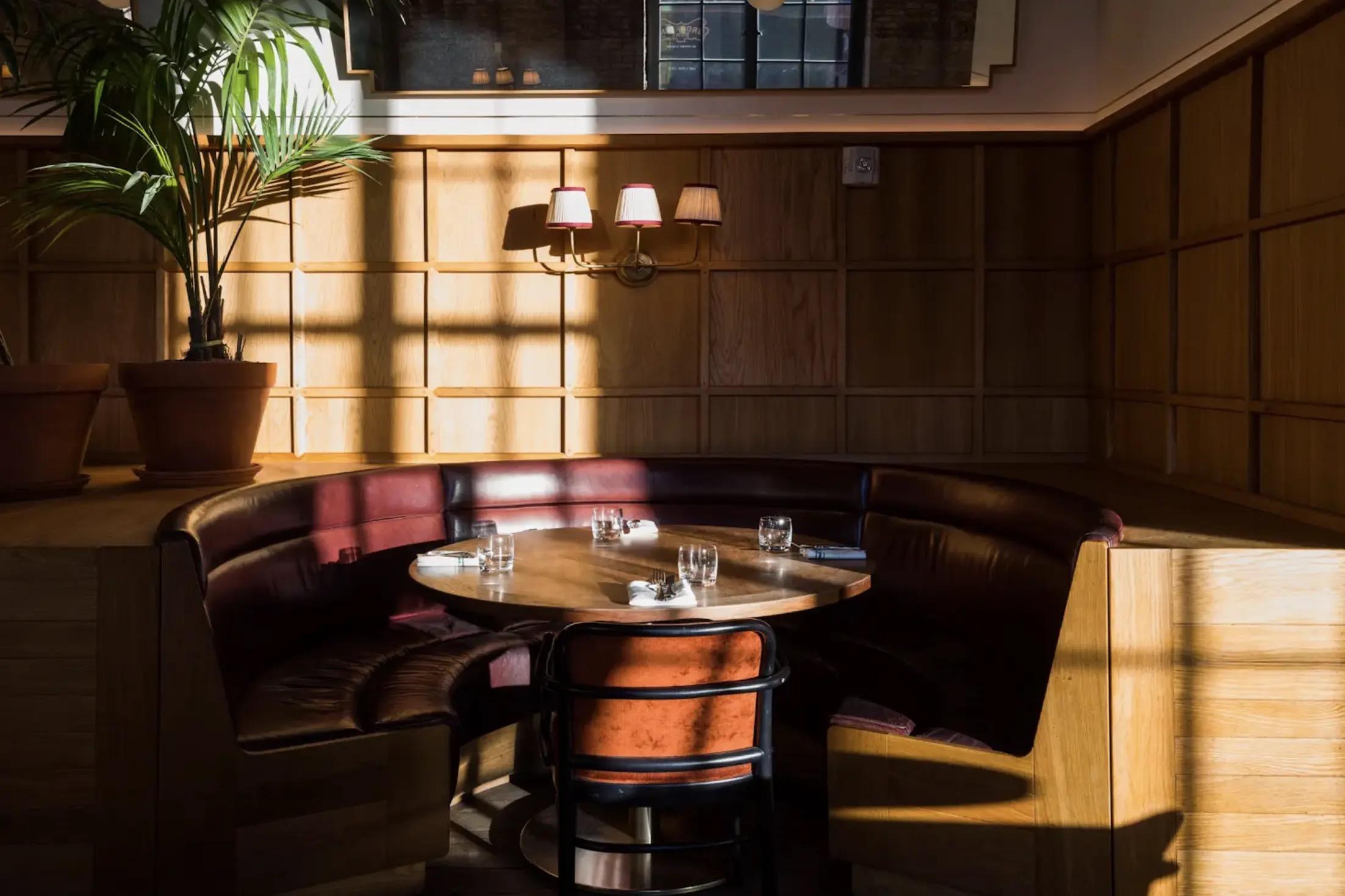 A curved red leather booth around a round wooden table set with glassware, in a window lit dining room with potted palms and wall sconces.