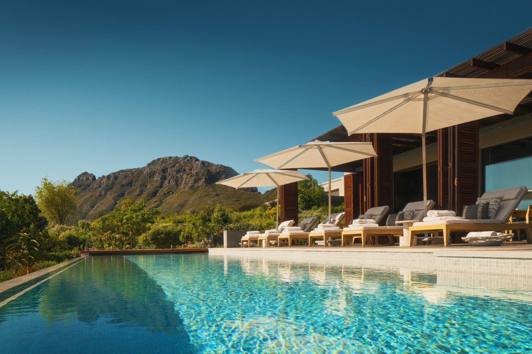 An infinity pool with shaded loungers along one side looks out to a rocky mountain under a clear sky.
