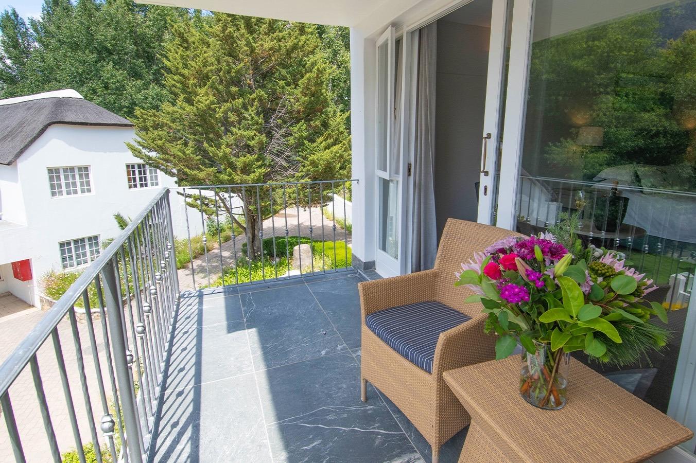 A balcony with two wicker chairs and a vase of flowers, looking down on a thatched white cottage and a garden path.