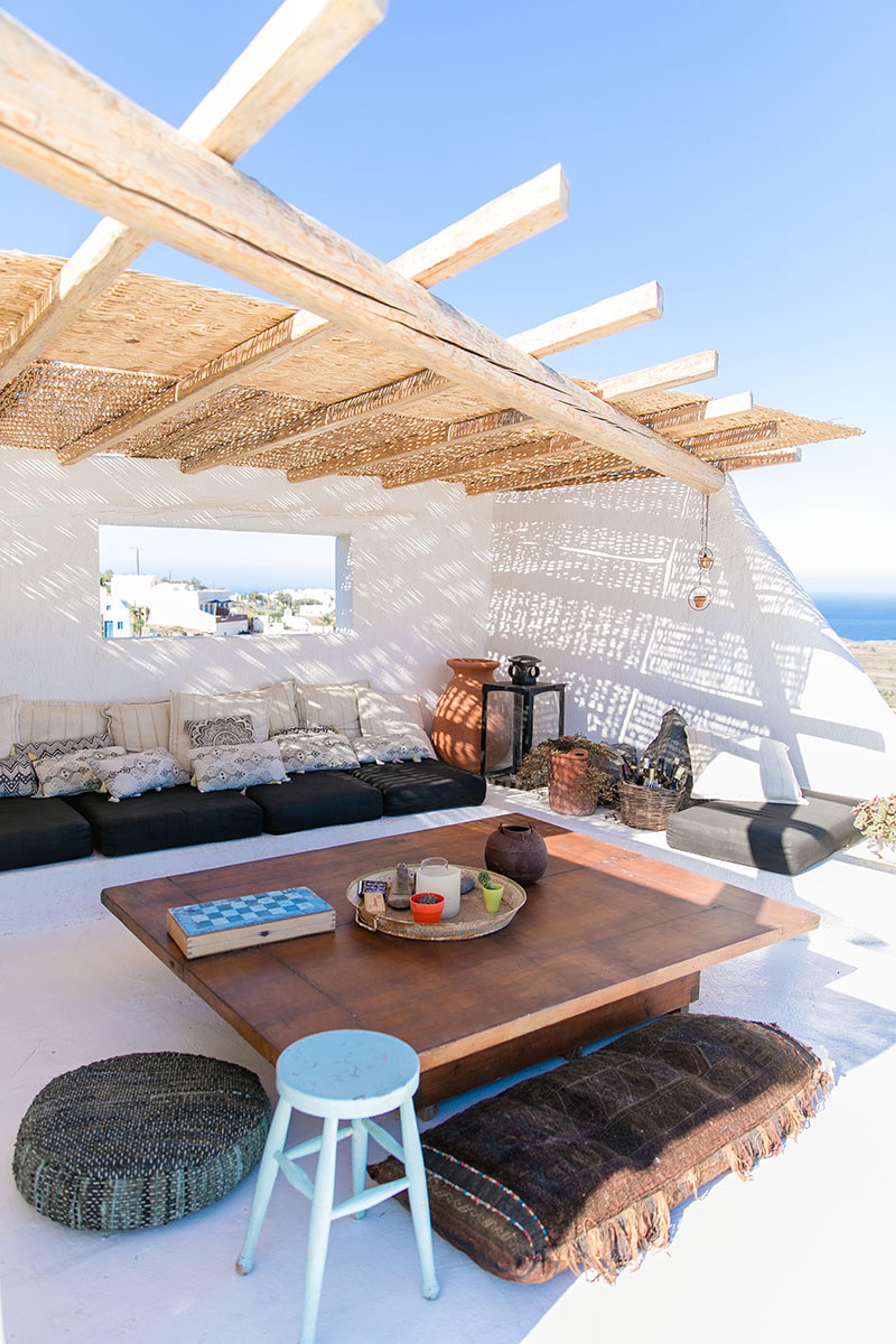 A shaded outdoor lounge under a timber pergola with woven reed, with a low dark sofa piled with patterned cushions, a wooden table, and a rug on the floor.