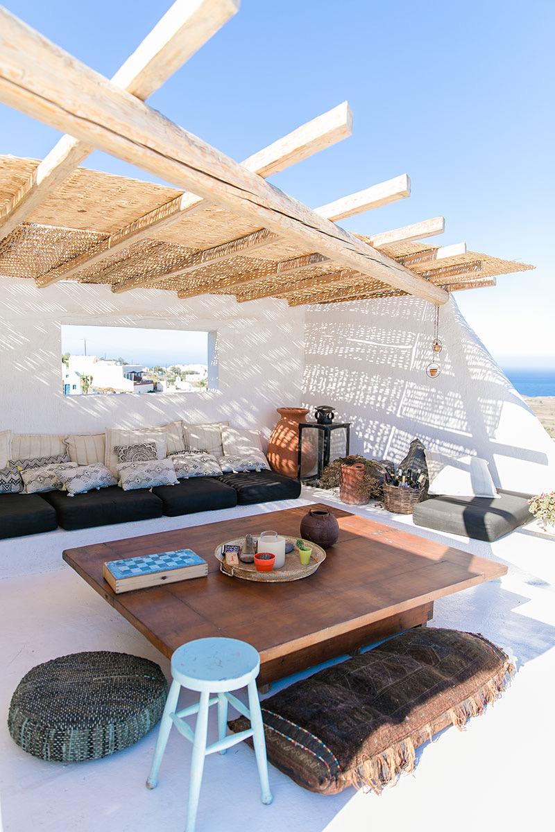 A shaded outdoor lounge under a timber pergola with woven reed, with a low dark sofa piled with patterned cushions, a wooden table, and a rug on the floor.