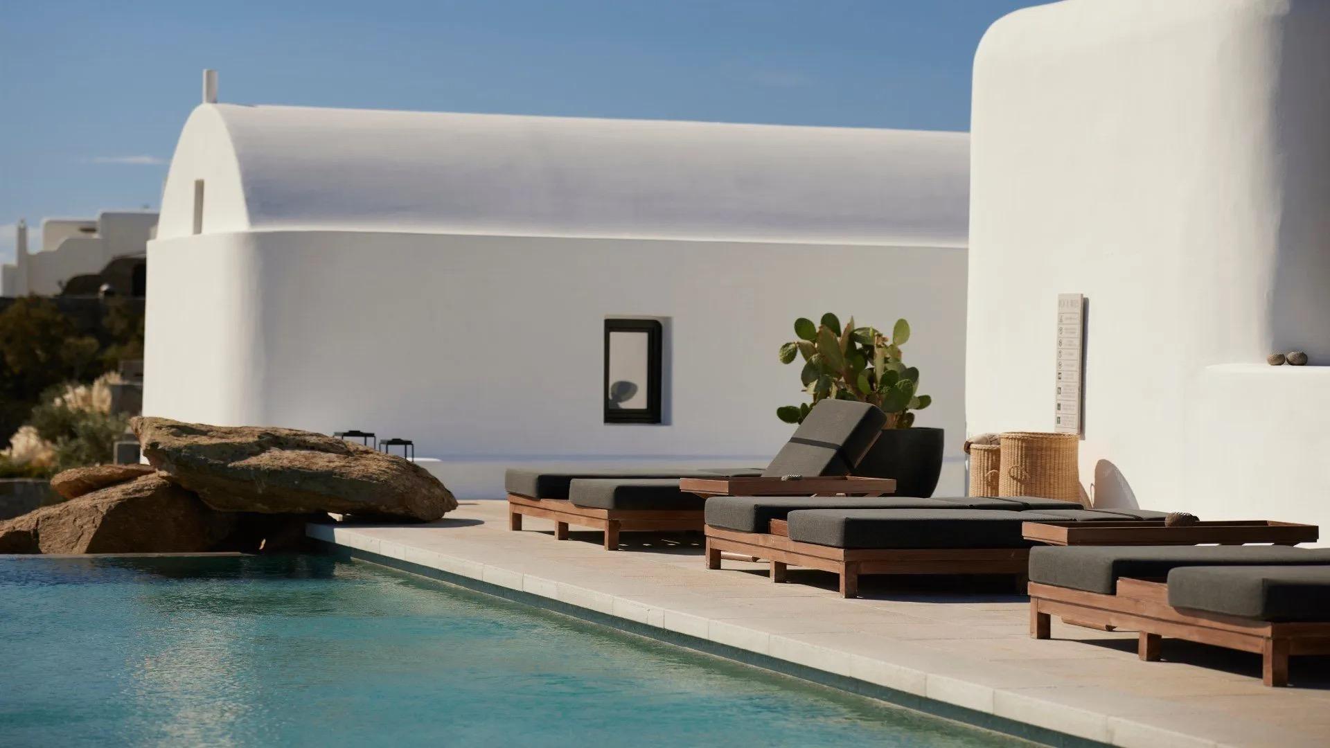 A domed white building beside a pool with dark sunloungers, a large boulder and a cactus at the water's edge.