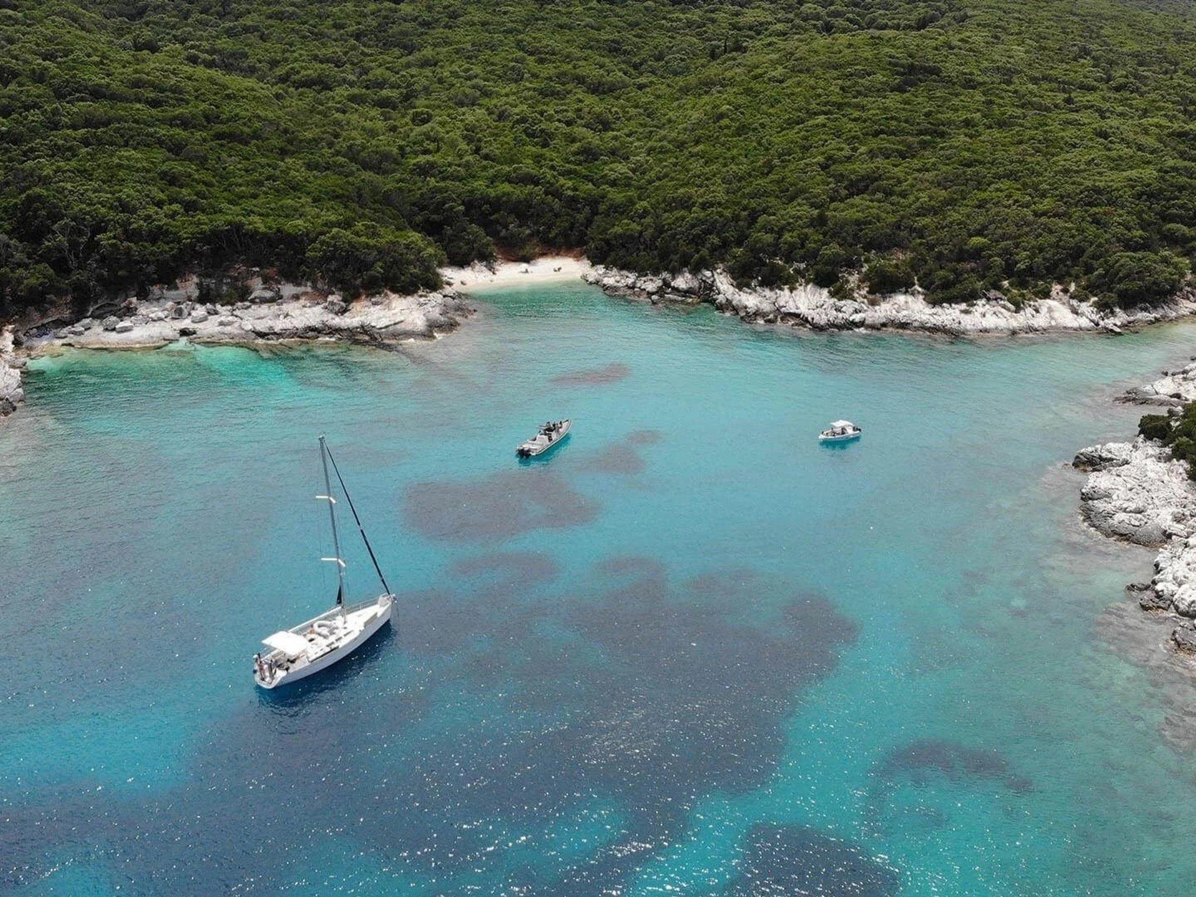 Boats anchored in a turquoise cove below pine-covered hills and white limestone, near Fiskardo
