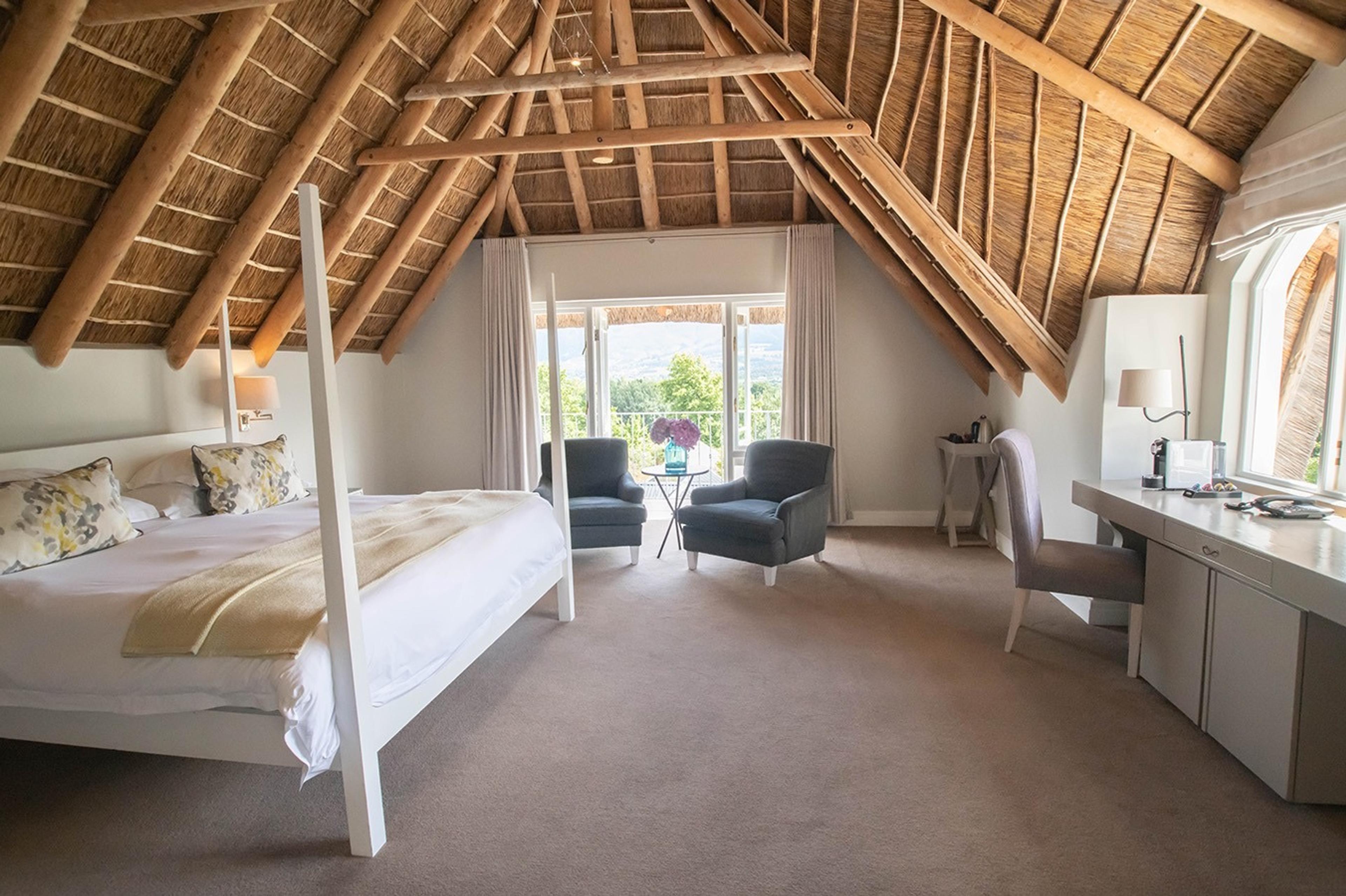 A bedroom with a white four-poster bed under an exposed thatched roof, with armchairs and tall windows onto trees and mountains.