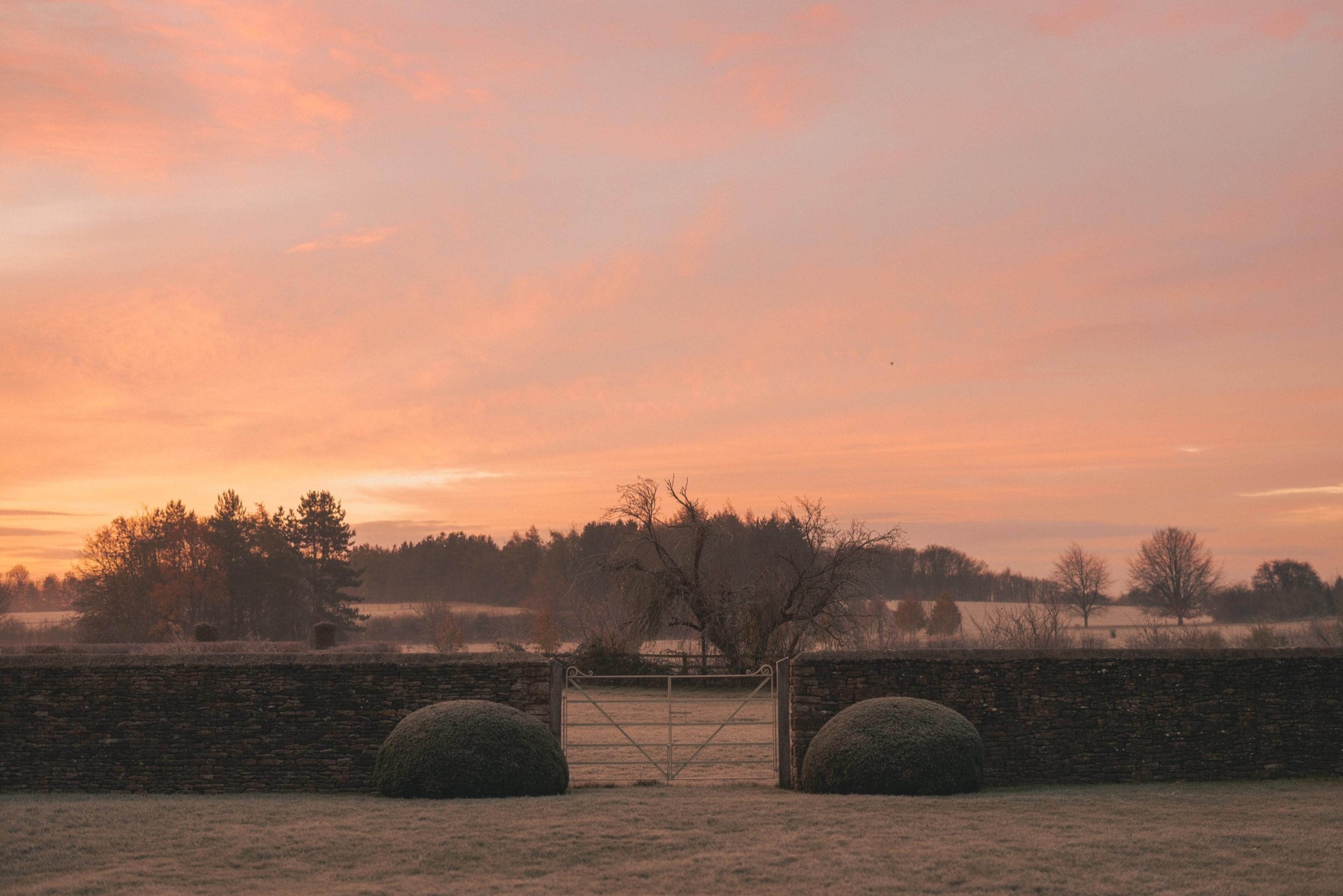 A stone wall and metal field gate under a pink and orange dawn sky, with trees and open fields beyond.