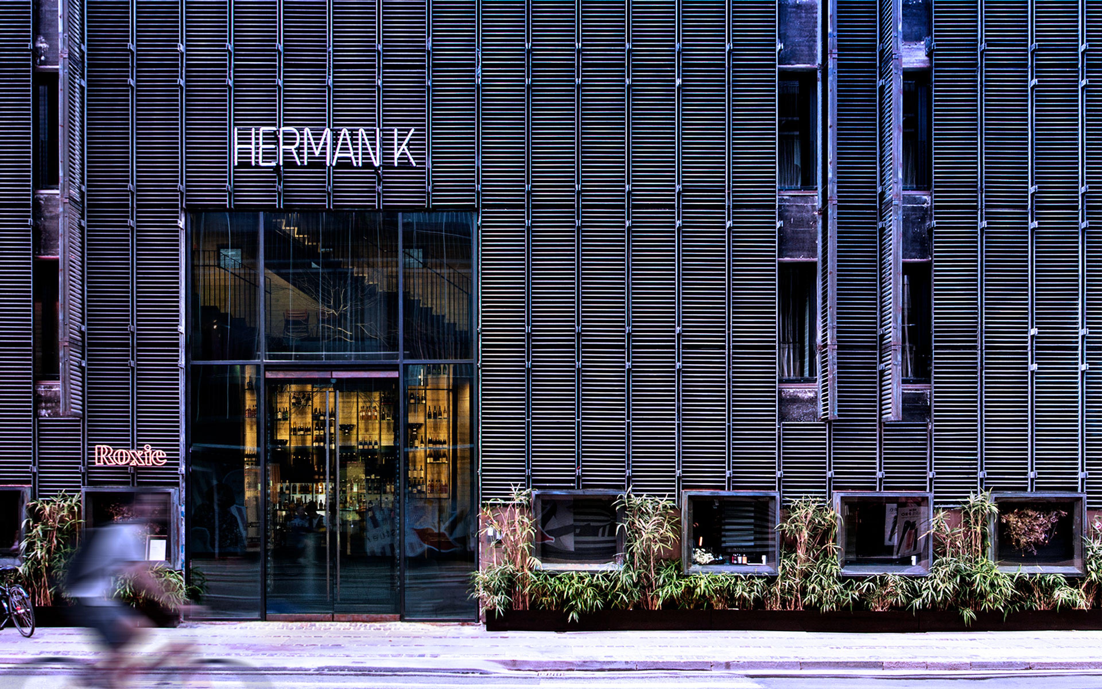 The building's facade at night, lit from within, with a glass entrance and the hotel's name in neon lettering above the door, and a cyclist passing by in motion blur.