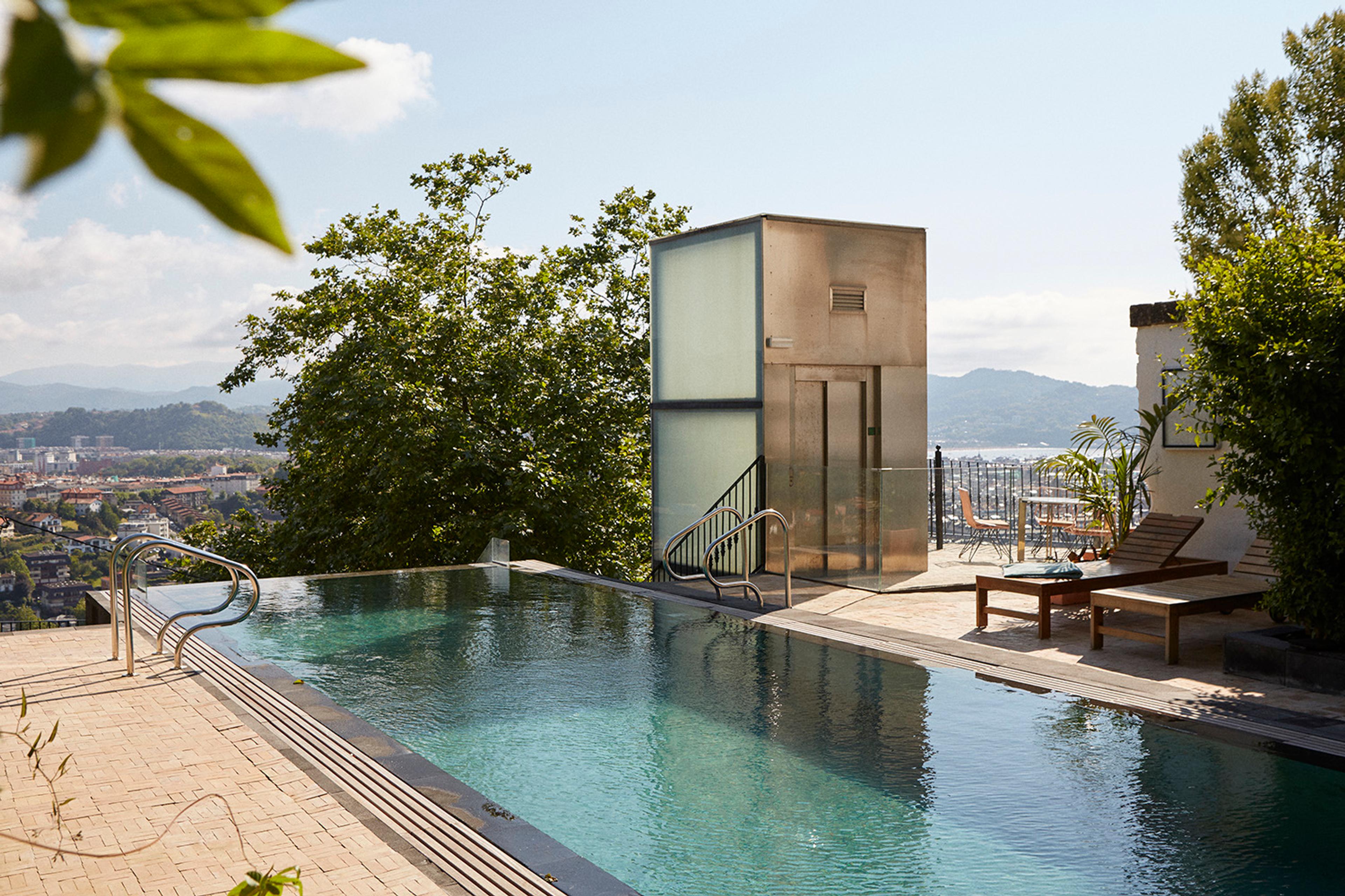 an infinity pool beside a glazed lift shaft, with sun loungers, a table and chairs under trees and the town and hills beyond