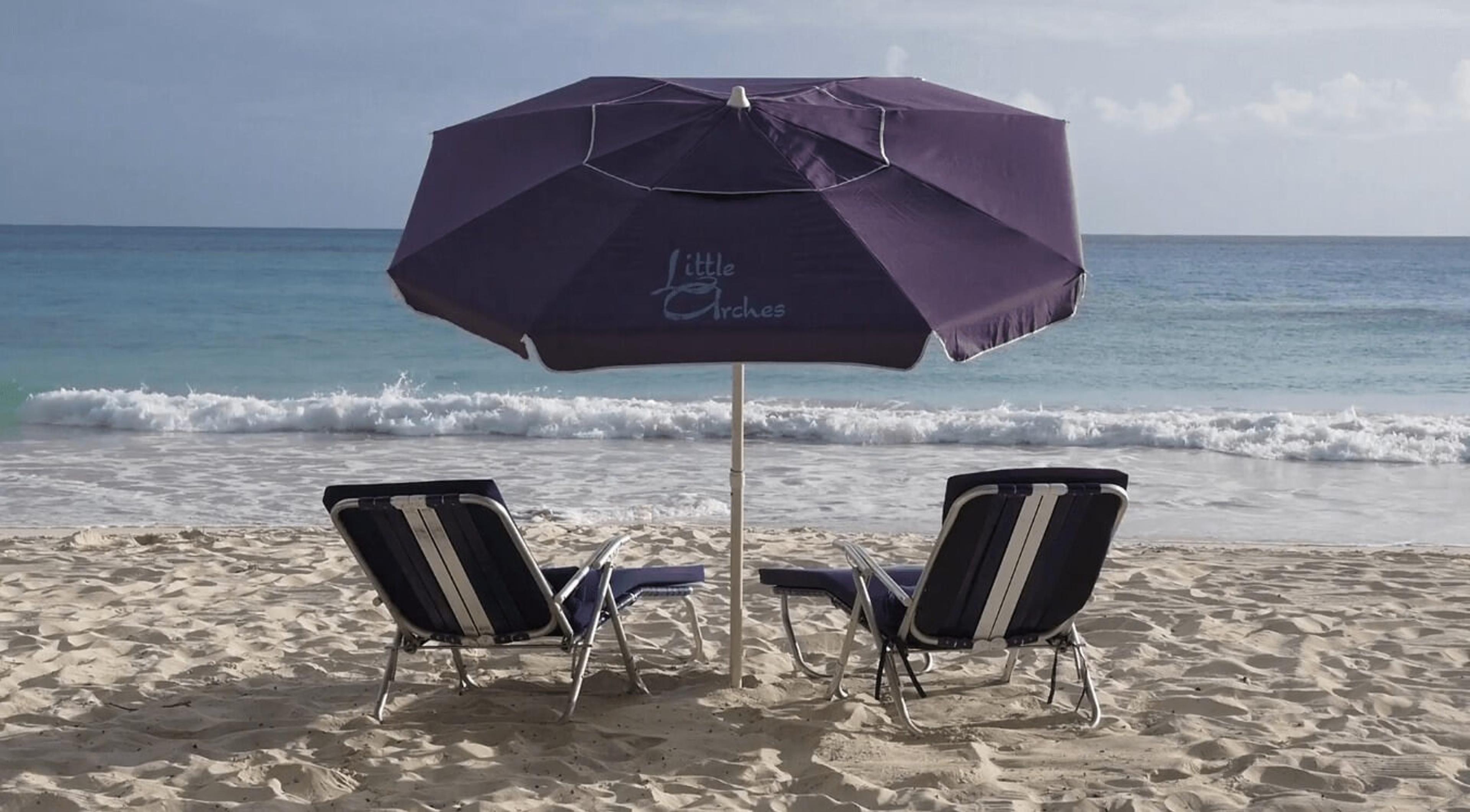 Two empty beach loungers under a purple umbrella printed with the Little Arches name, facing the sea.