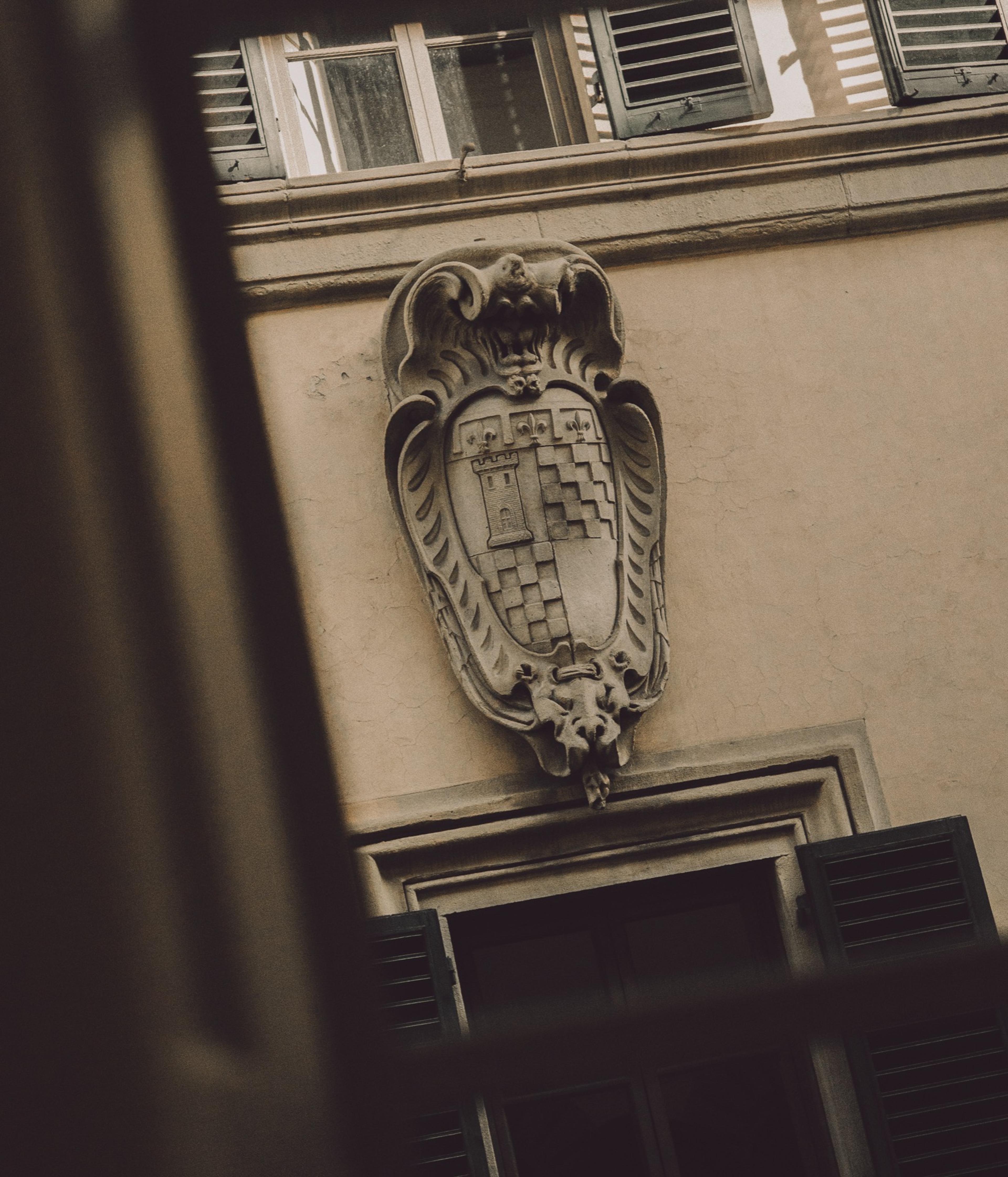 A carved stone coat of arms with a tower and shield sits above a shuttered window on the building's sandstone facade.