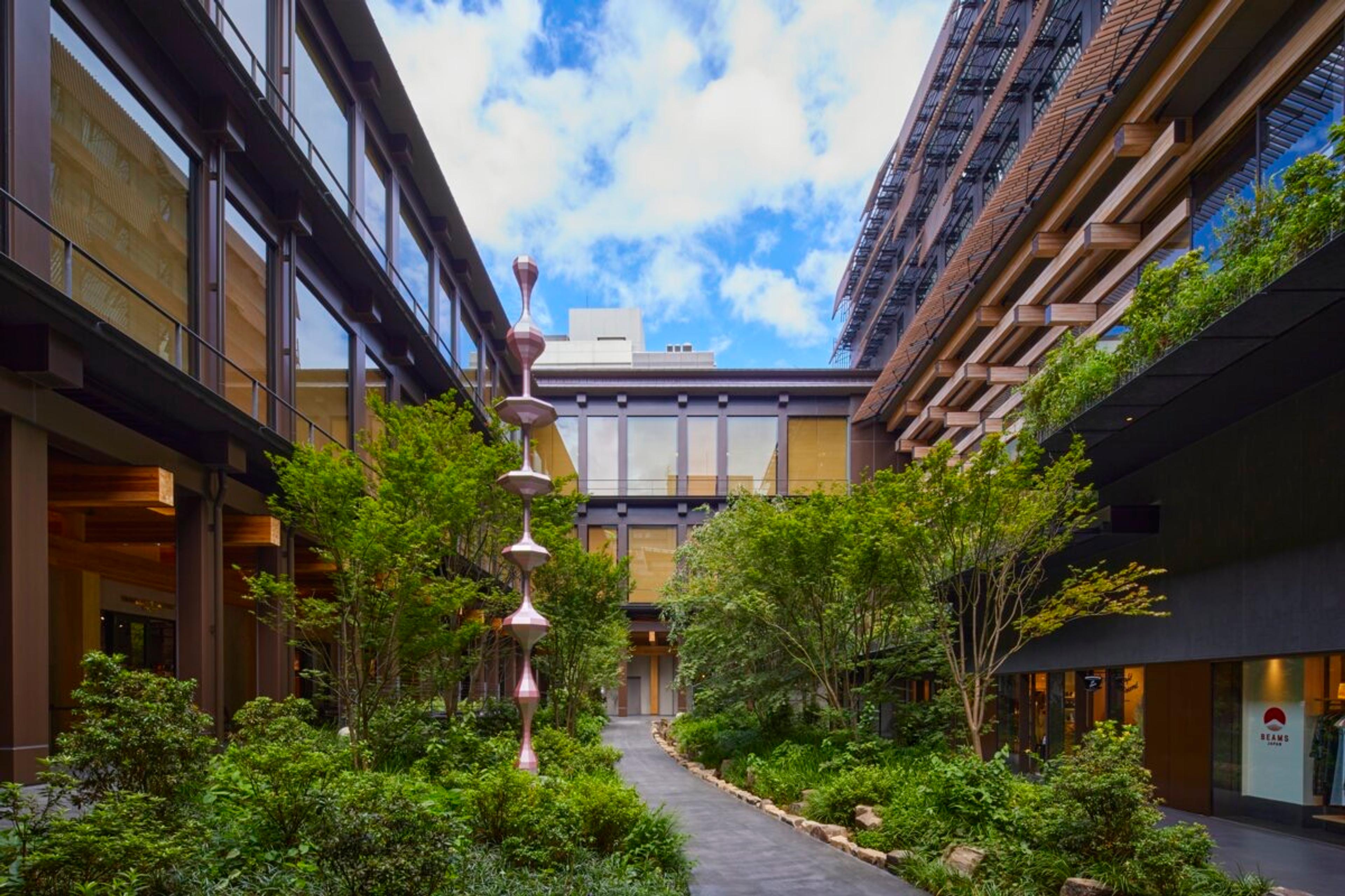A courtyard between two wings of the hotel with a stone path, planted beds, and a tall sculpture of stacked pink forms, under a blue sky with clouds.