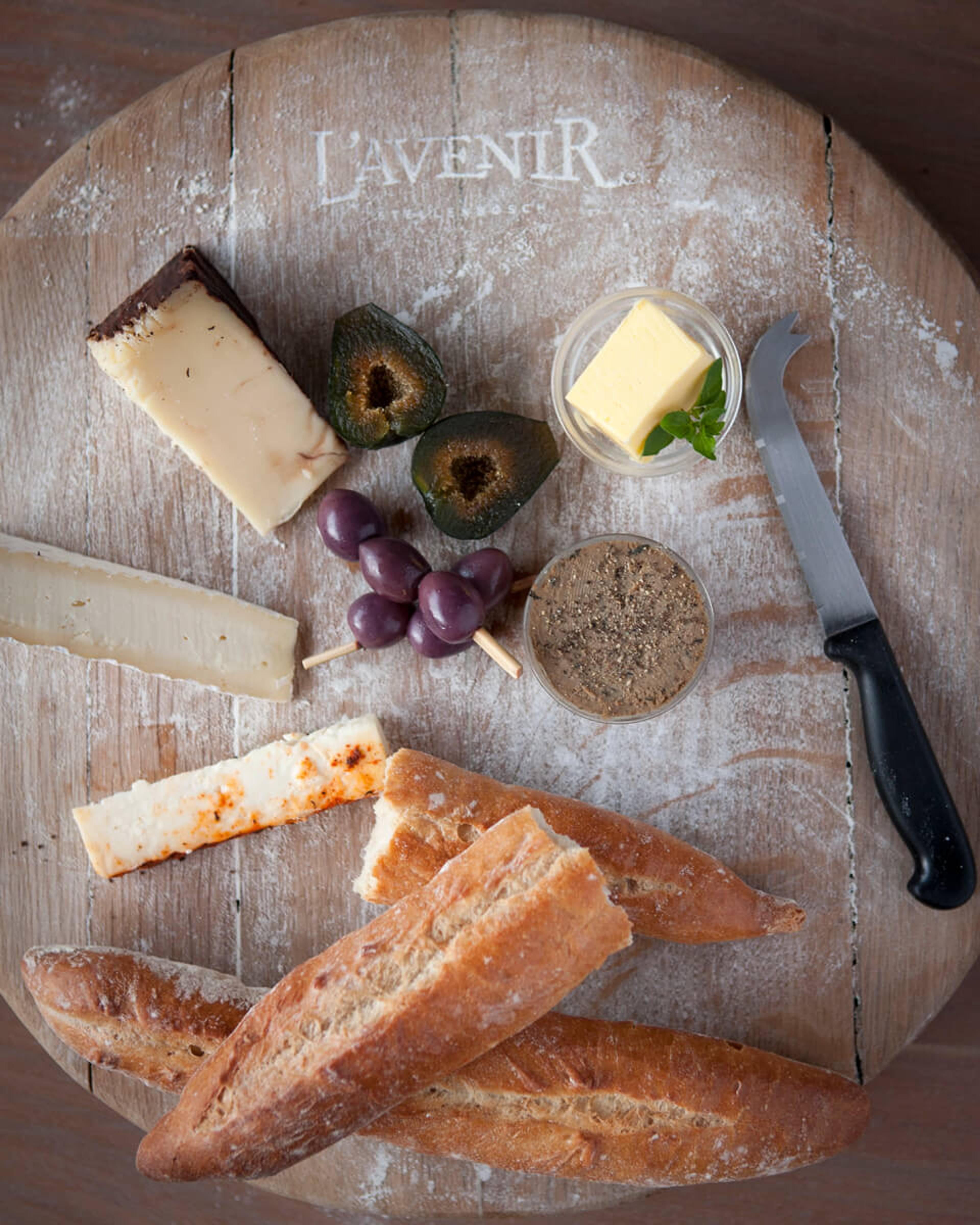 A round wooden board with cheeses, olives on sticks, a pat of butter, a dish of pâté, two baguettes and a cheese knife.