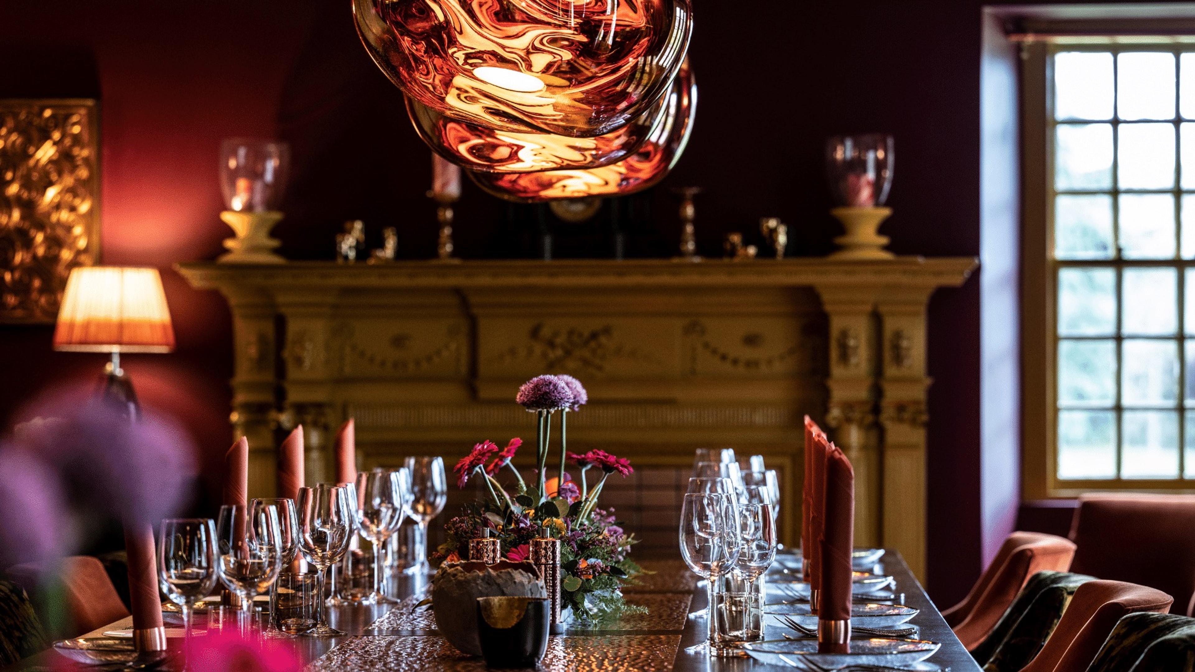 A dining table laid with wine glasses, red napkins and a flower arrangement, beneath a copper pendant light and in front of a carved mantelpiece