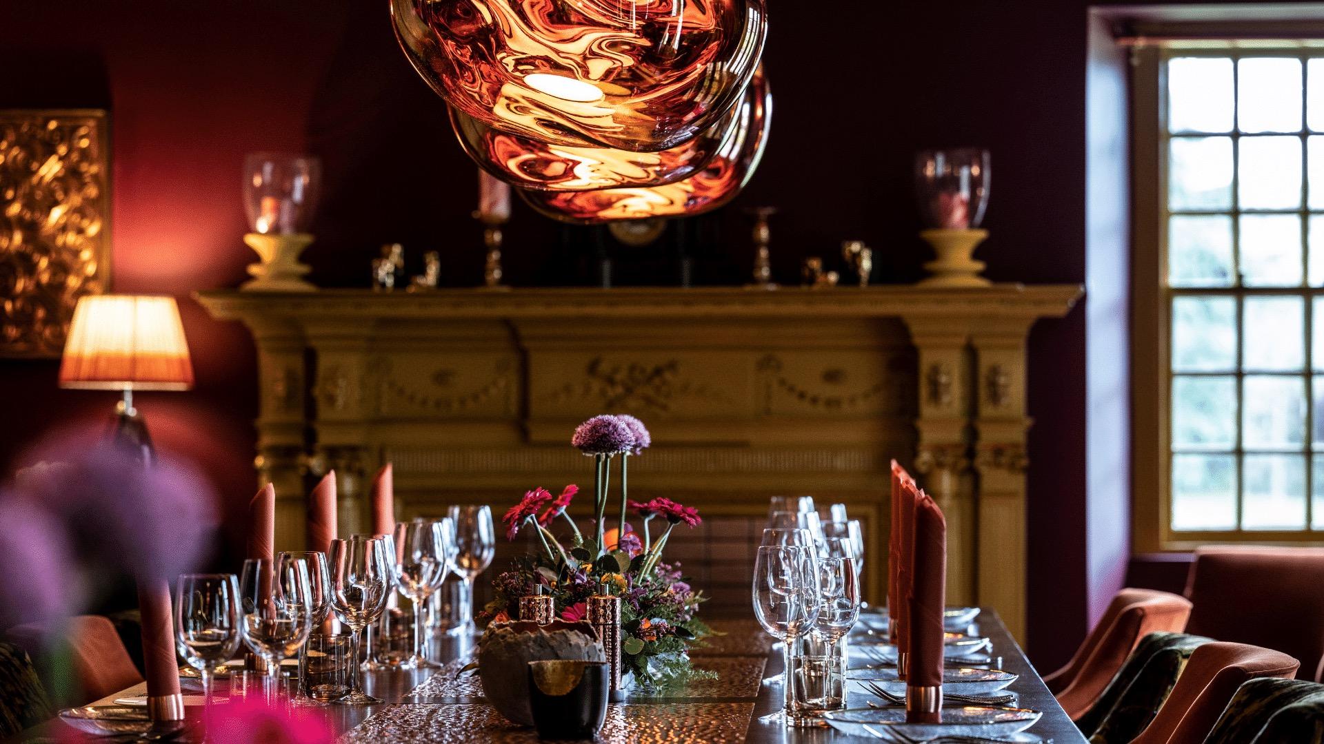 A dining table laid with wine glasses, red napkins and a flower arrangement, beneath a copper pendant light and in front of a carved mantelpiece