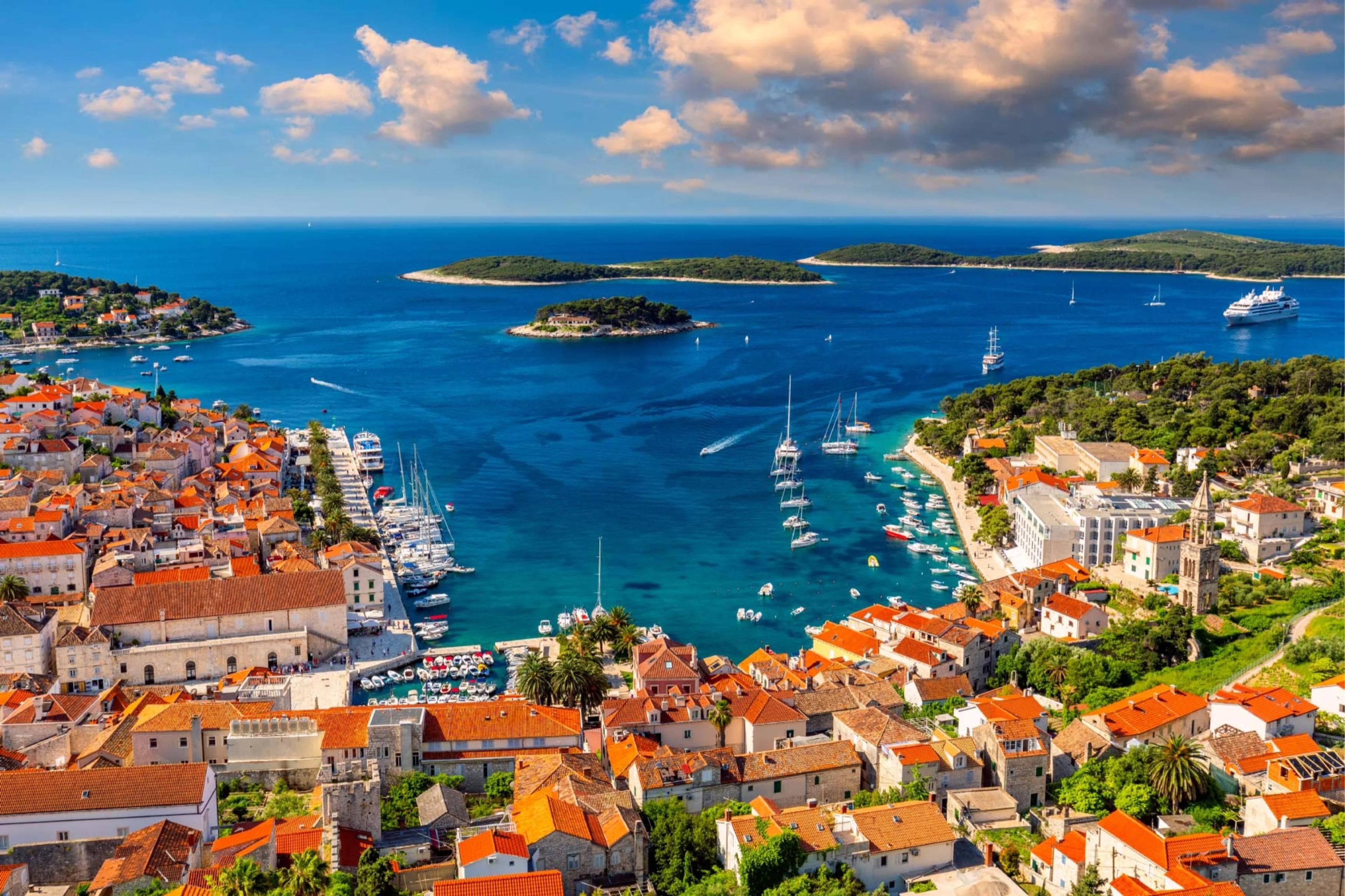 An aerial view of a harbour town with orange-roofed buildings, moored yachts, and wooded islands beyond.