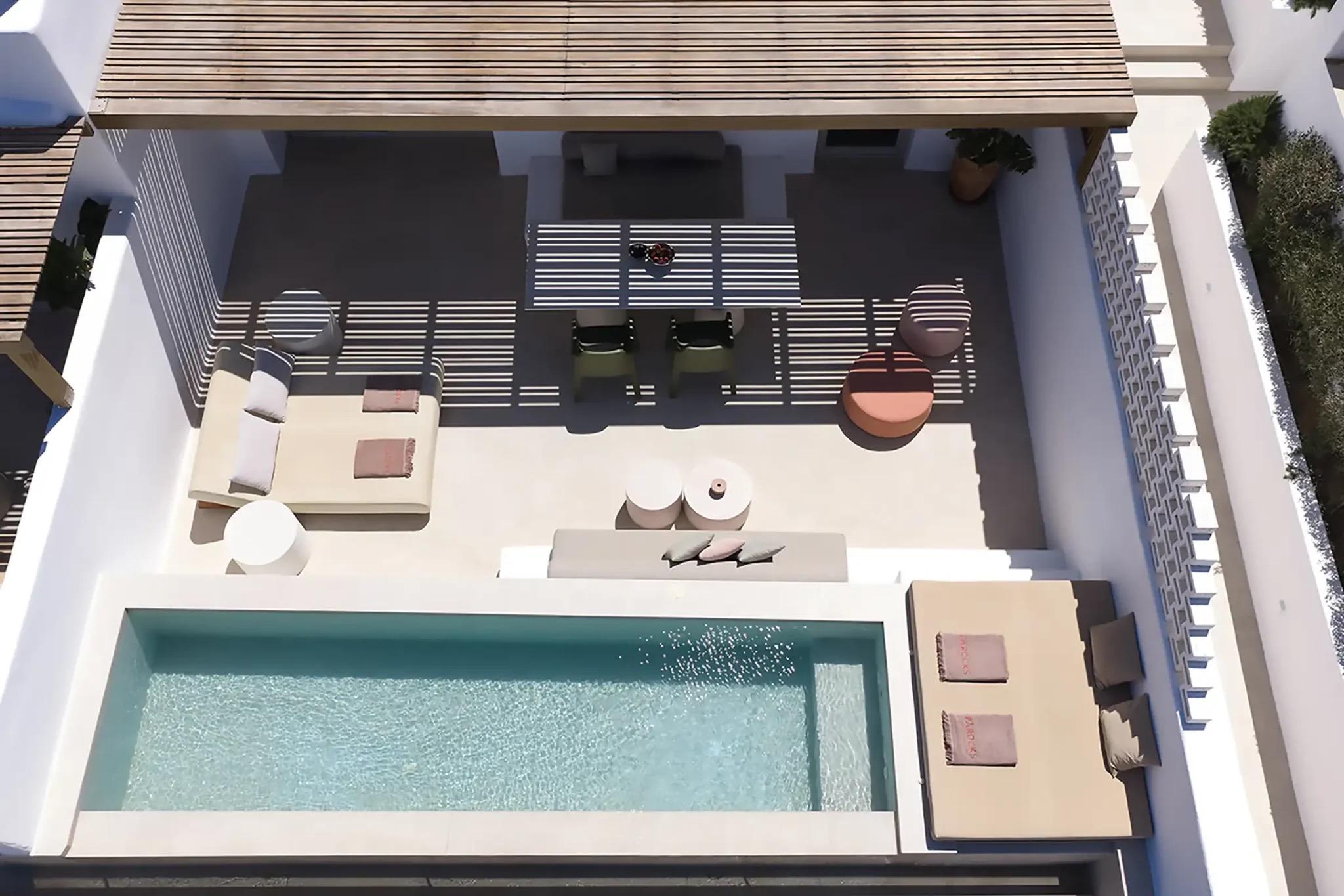 An aerial view looking down into a narrow infinity-edged plunge pool on a private terrace with a pergola, sunbeds and a dining table.