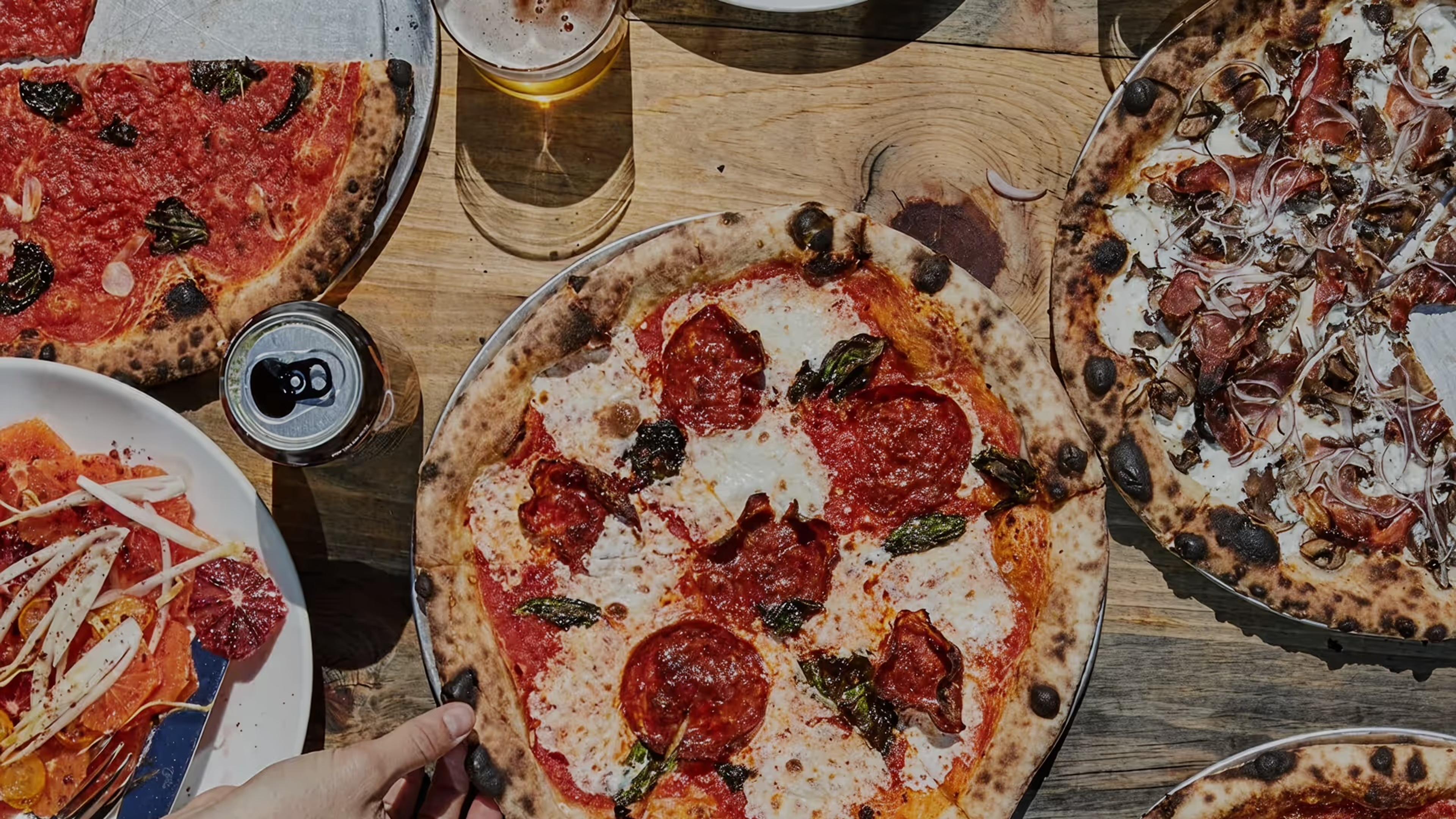 A pepperoni pizza on a wooden table among other pizzas and cans, seen from above