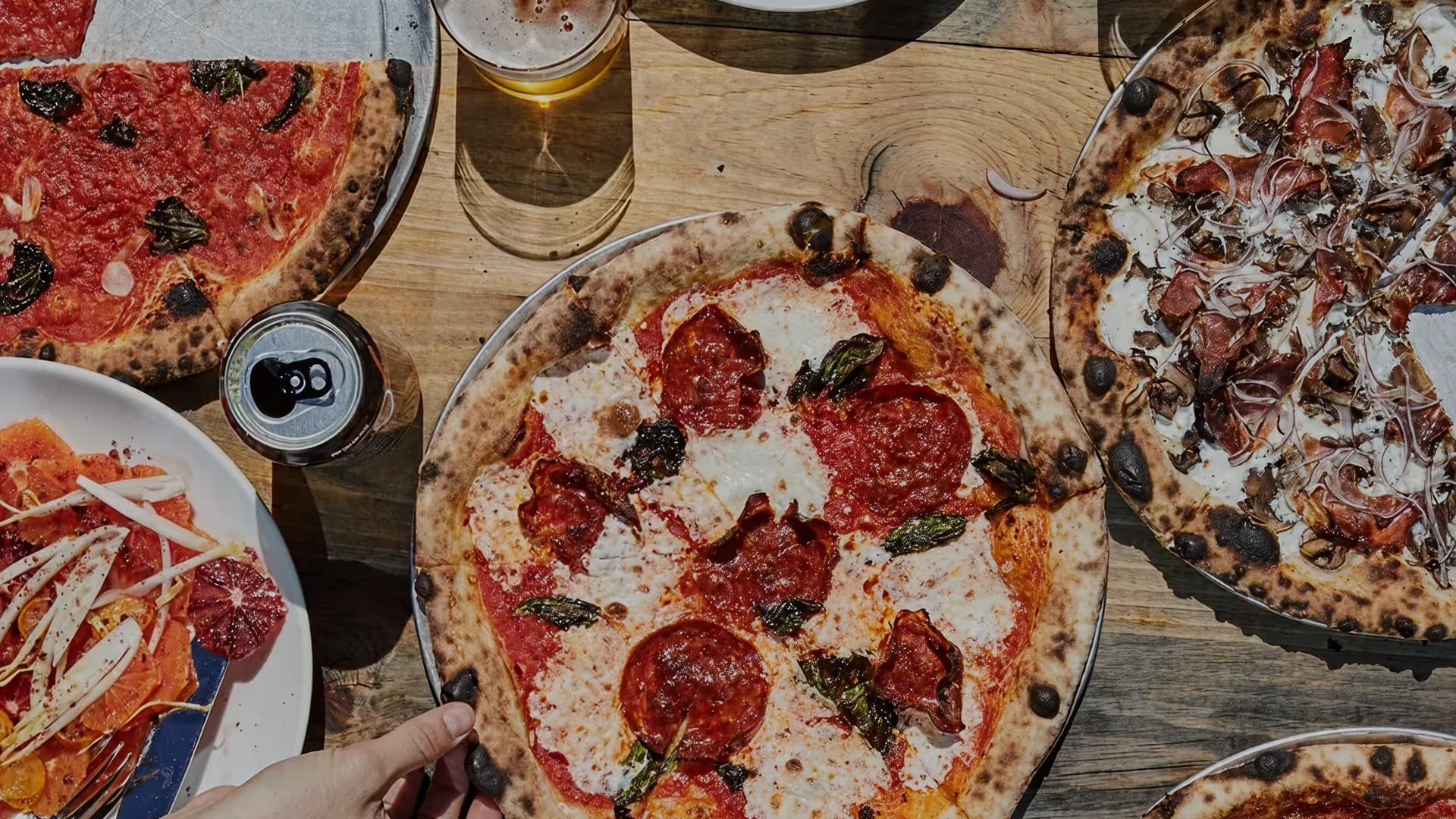 A pepperoni pizza on a wooden table among other pizzas and cans, seen from above