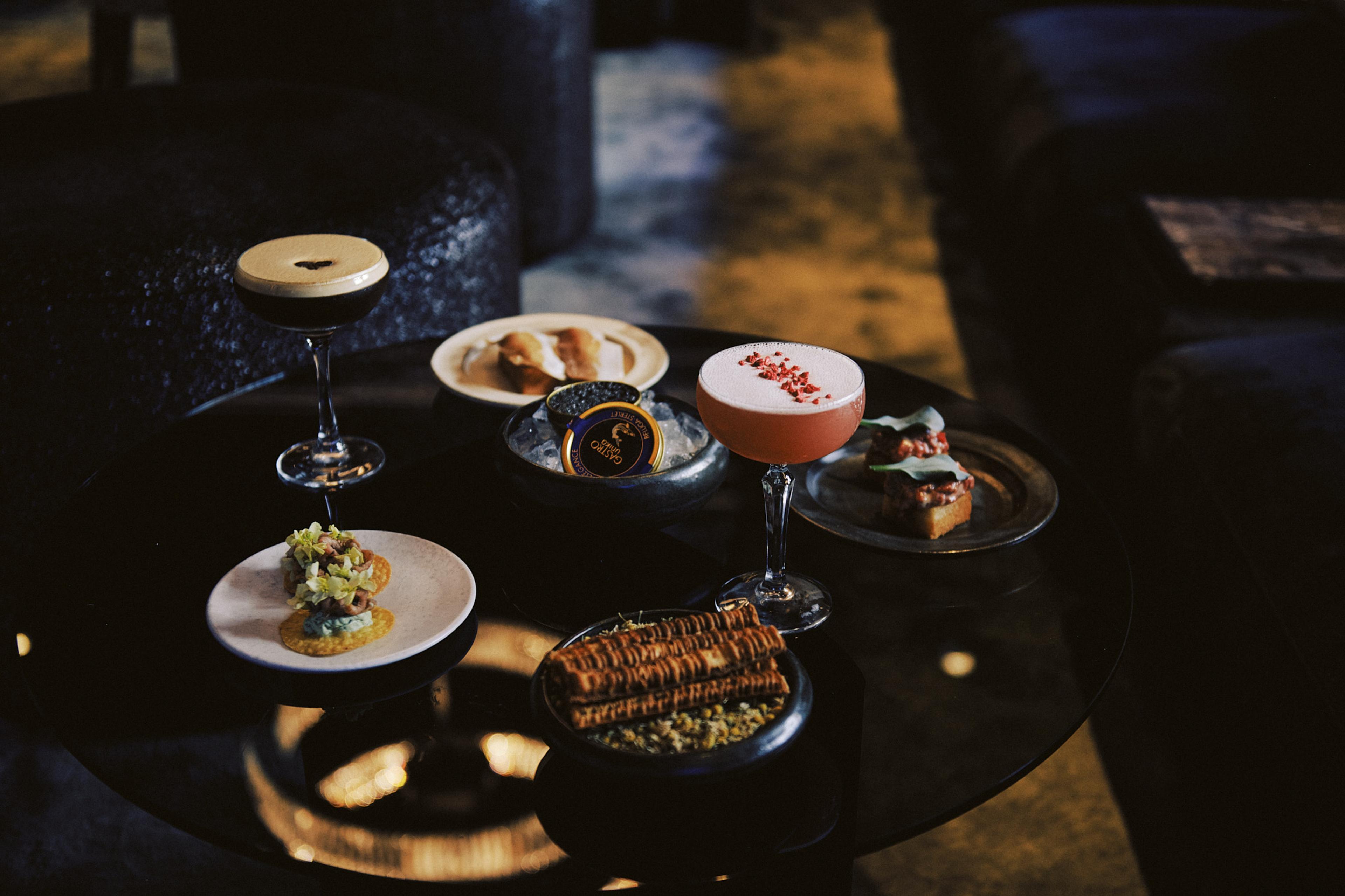 A low table holding cocktails and small plates of food, including toast, caviar and bruschetta, in a dimly lit room.