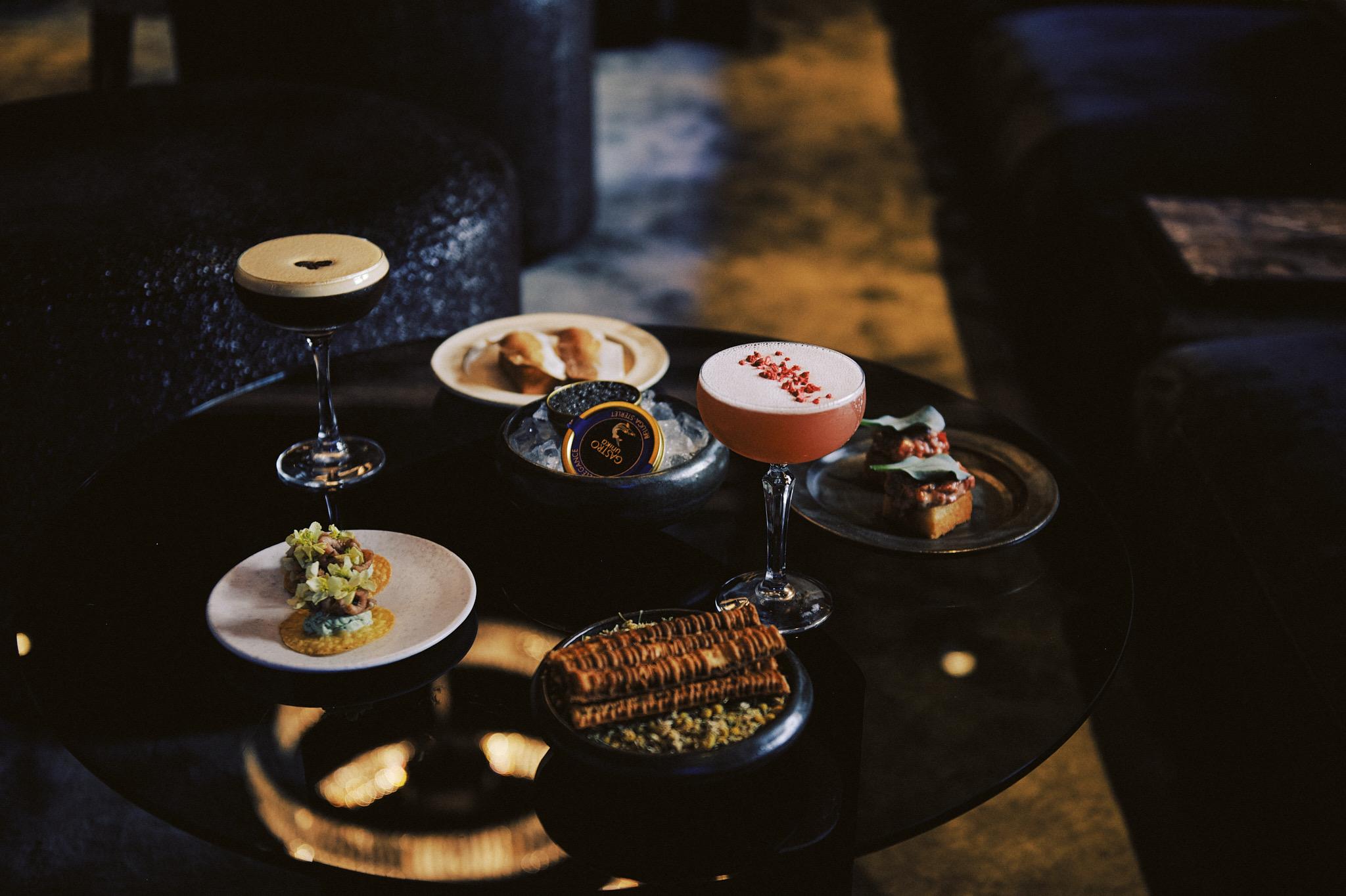 A low table holding cocktails and small plates of food, including toast, caviar and bruschetta, in a dimly lit room.