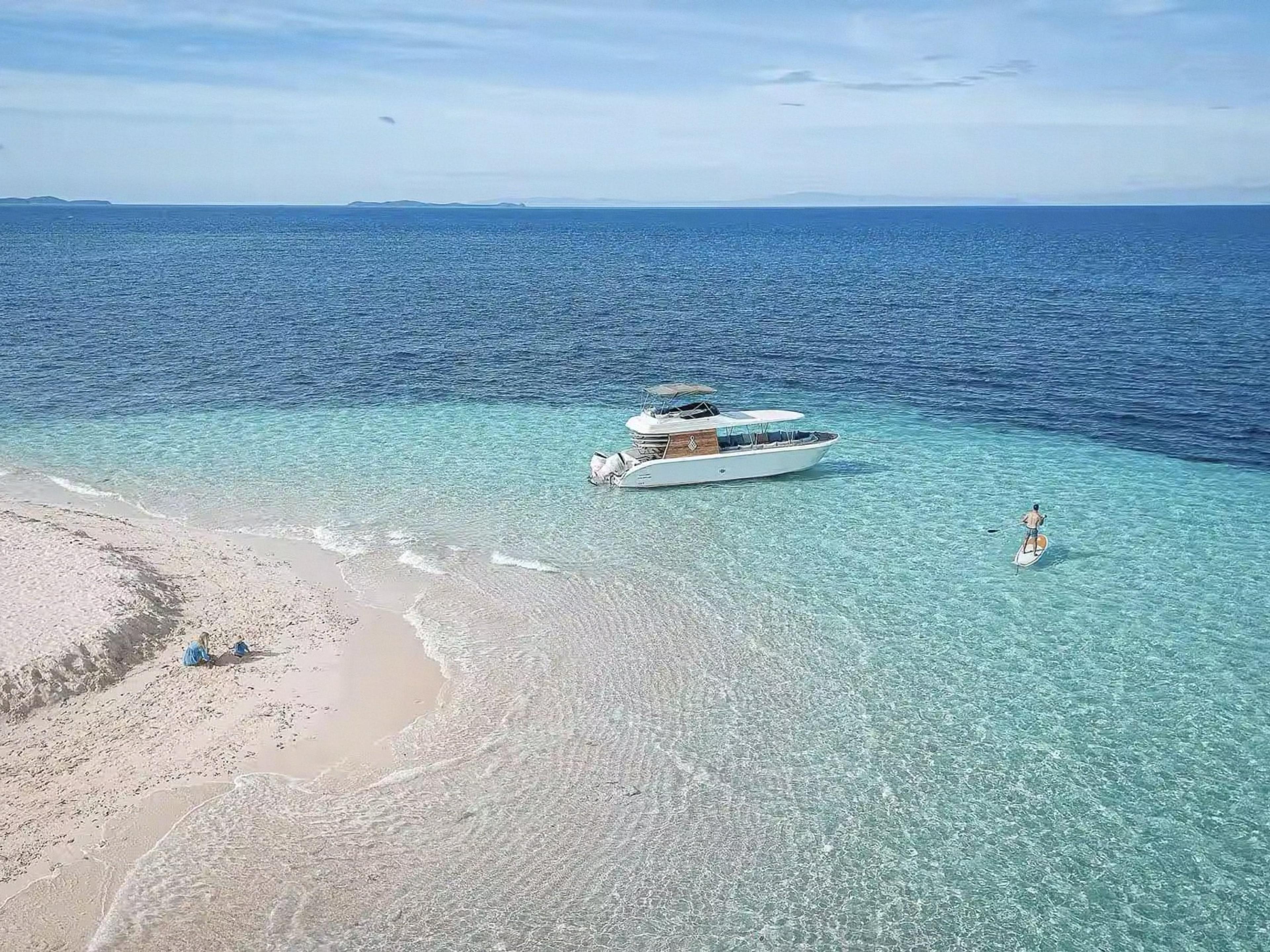 A speedboat anchored off a white sandbar in clear turquoise water, a paddleboarder beside it