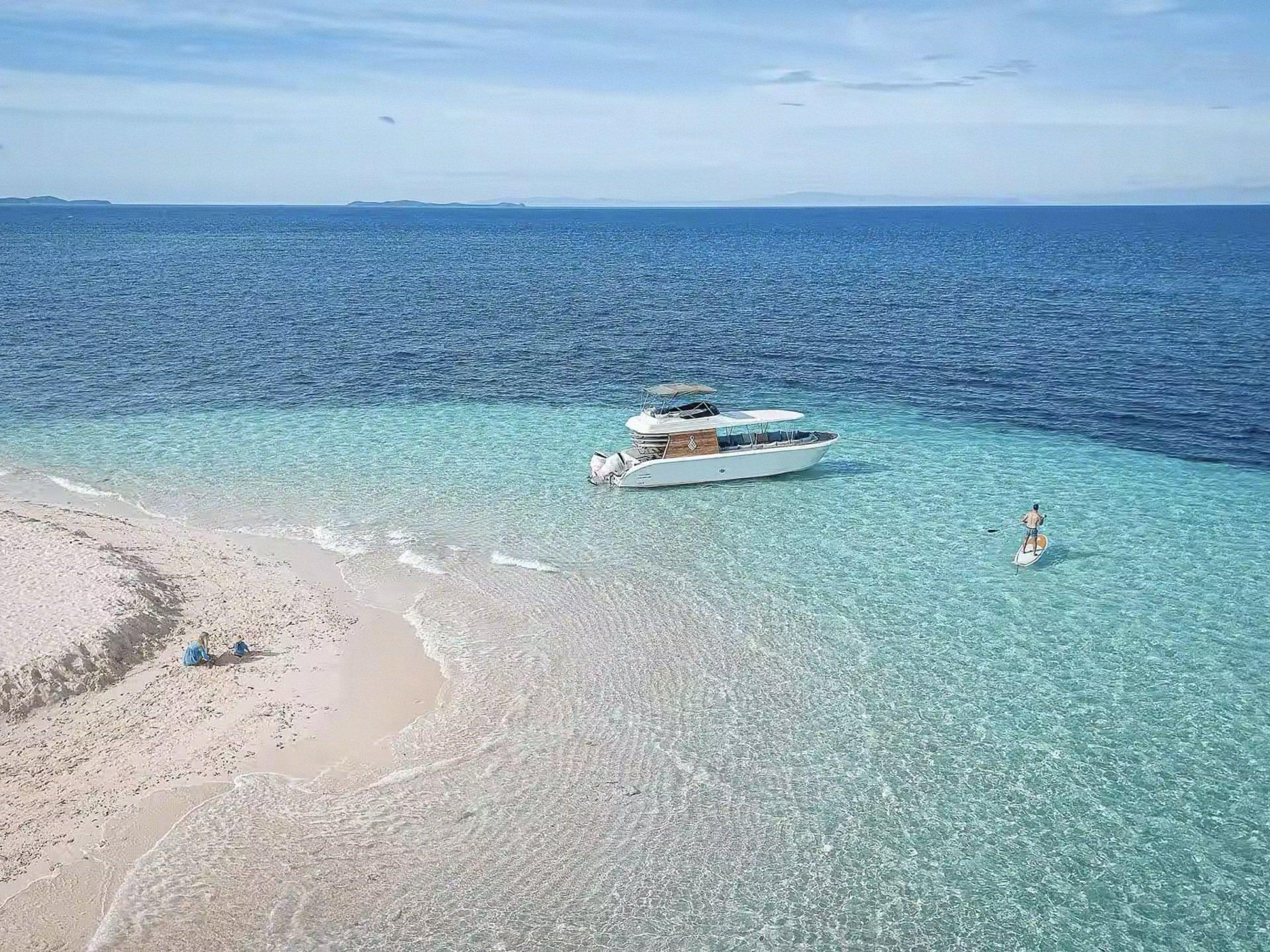 A speedboat anchored off a white sandbar in clear turquoise water, a paddleboarder beside it