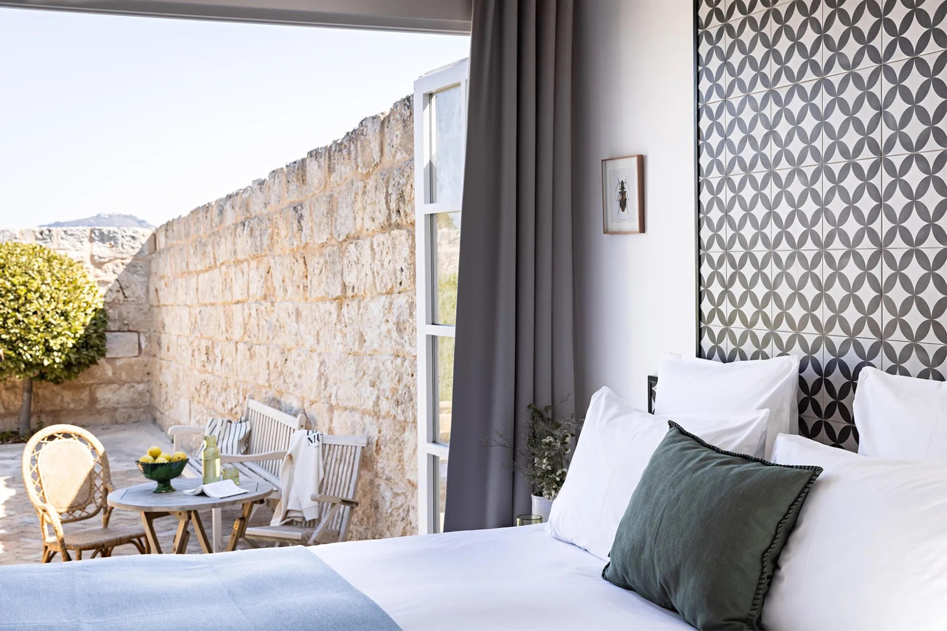 a bedroom with a patterned black and white tiled headboard and a bed made up in white linen, its doors open onto a walled stone courtyard with rattan chairs and a table set with lemons